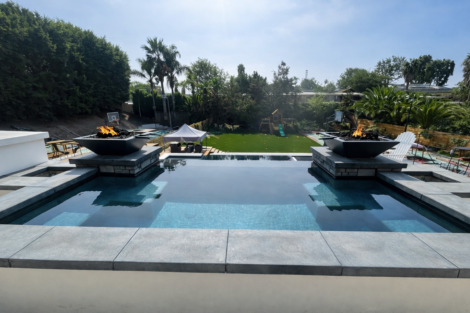 Backyard with a pool and two outdoor fire pits on a patio, overlooking a grassy area with trees and a playset.