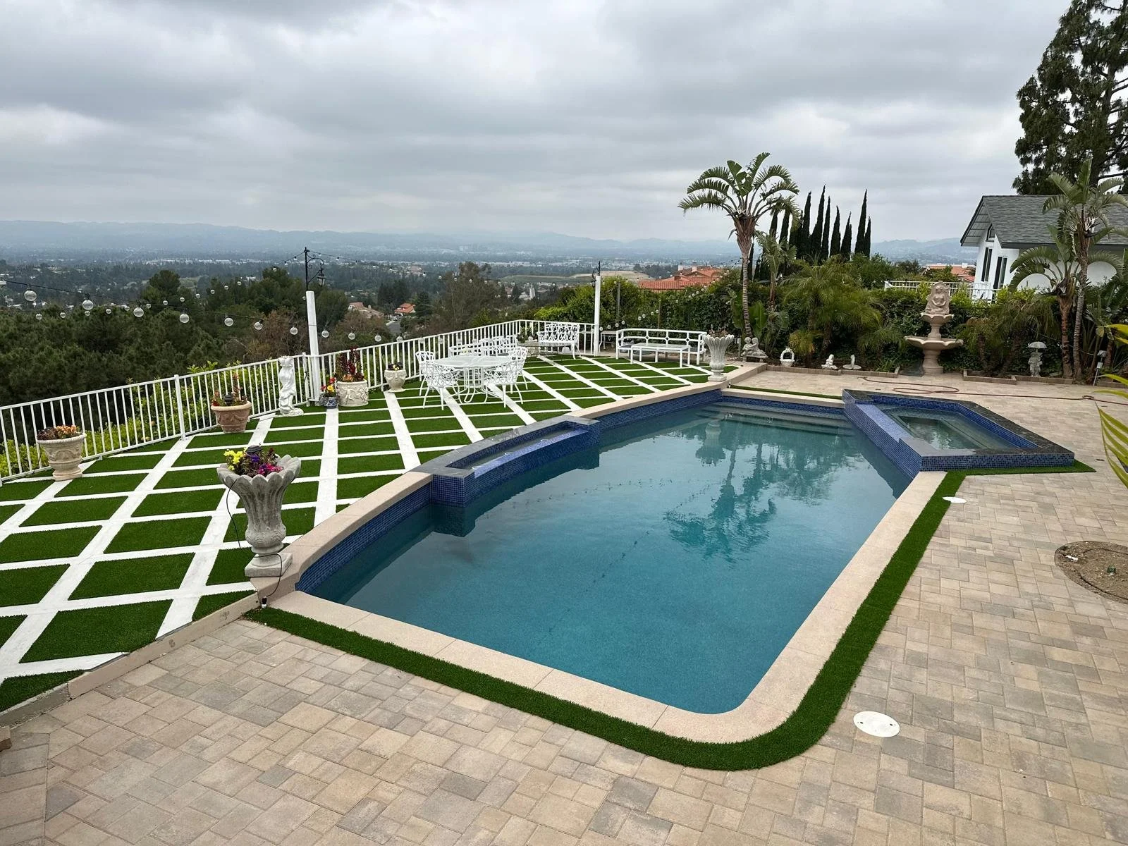 A backyard swimming pool with a hot tub surrounded by pavers and green turf, overlooking a scenic view of trees and city in the distance. The area is decorated with potted plants, white furniture, statues, and garden ornaments. Overcast sky