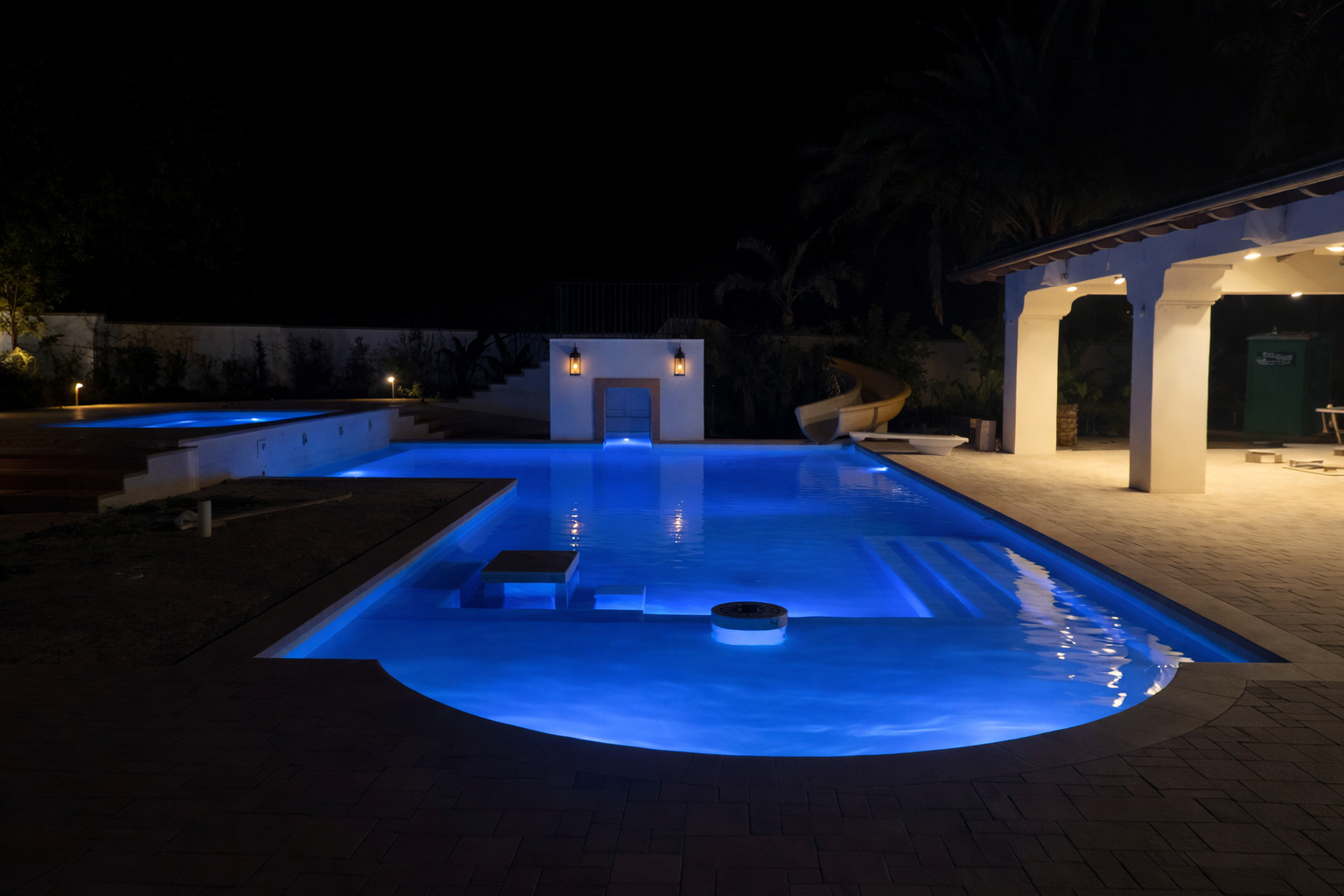 Night view of a lit swimming pool with blue underwater lights, a small slide at the far end, an outdoor lounge area with white pillars and a thatched roof, and surrounding greenery.