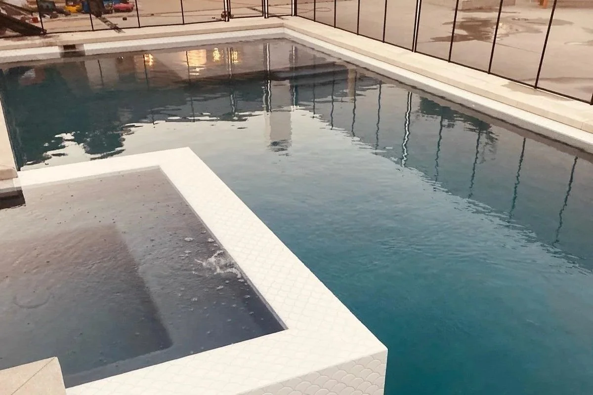 A swimming pool with a hot tub attached, surrounded by glass fencing, reflecting an evening sky.