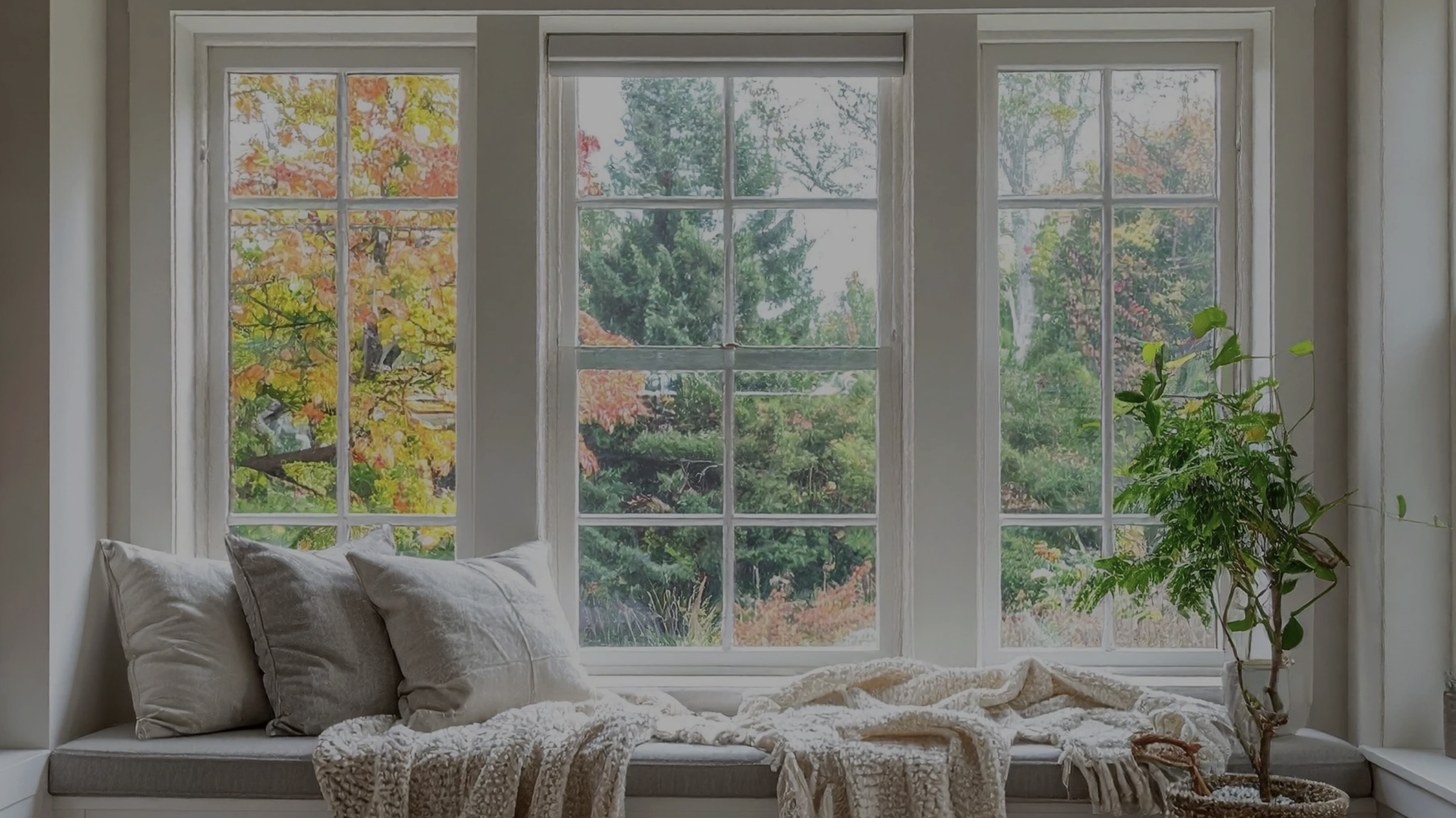 Cozy window seat with white grid windows overlooking colorful autumn trees, styled with neutral pillows, a soft throw, and a potted plant.