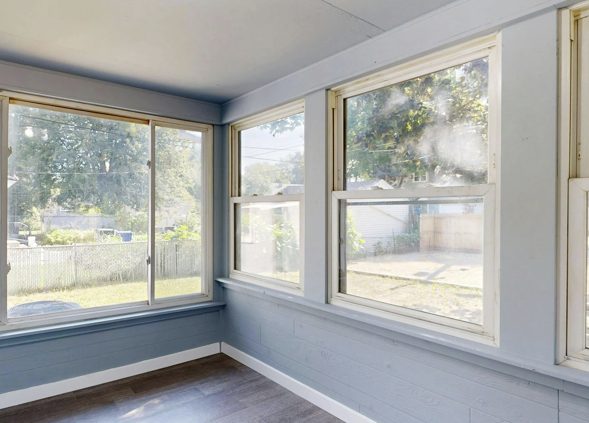 Bright sunroom with white-framed windows, blue lower wall paneling, and views of a fenced backyard filled with natural daylight.