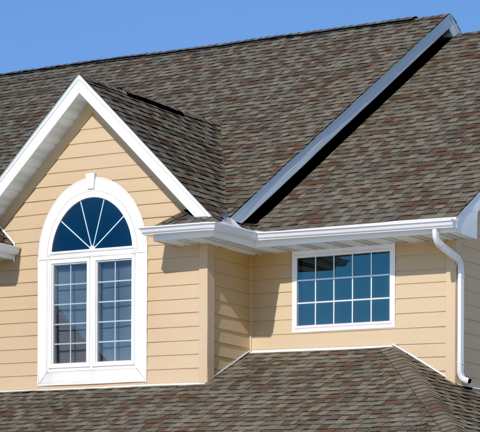 Close-up of a home exterior featuring beige siding, white-trim windows, and architectural asphalt shingles under a clear blue sky.