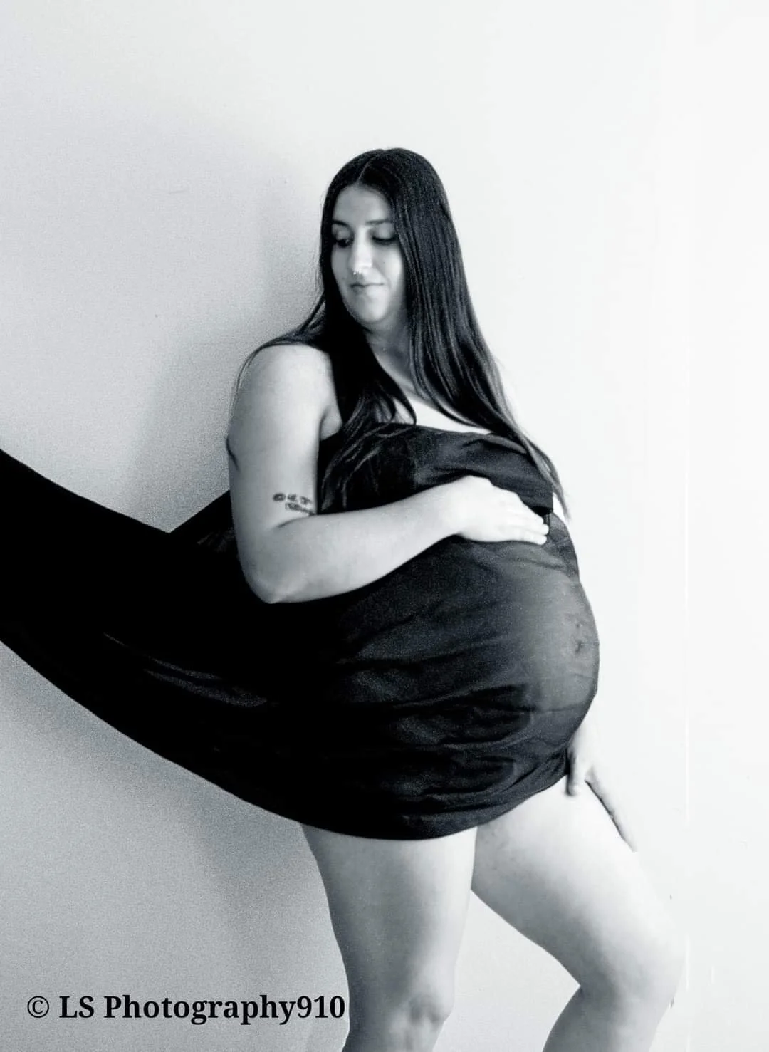 A timeless black-and-white maternity portrait capturing the quiet strength and beauty of motherhood. The expecting mother stands gracefully against a clean backdrop, draped in flowing fabric that gently supports and accentuates her baby bump. Her han