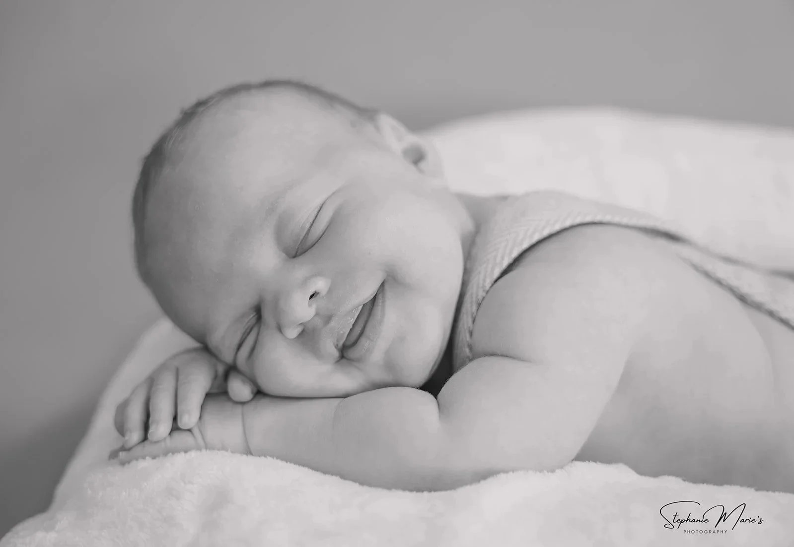 Black and white photo of a smiling baby sleeping on a soft blanket with arms folded under the head.