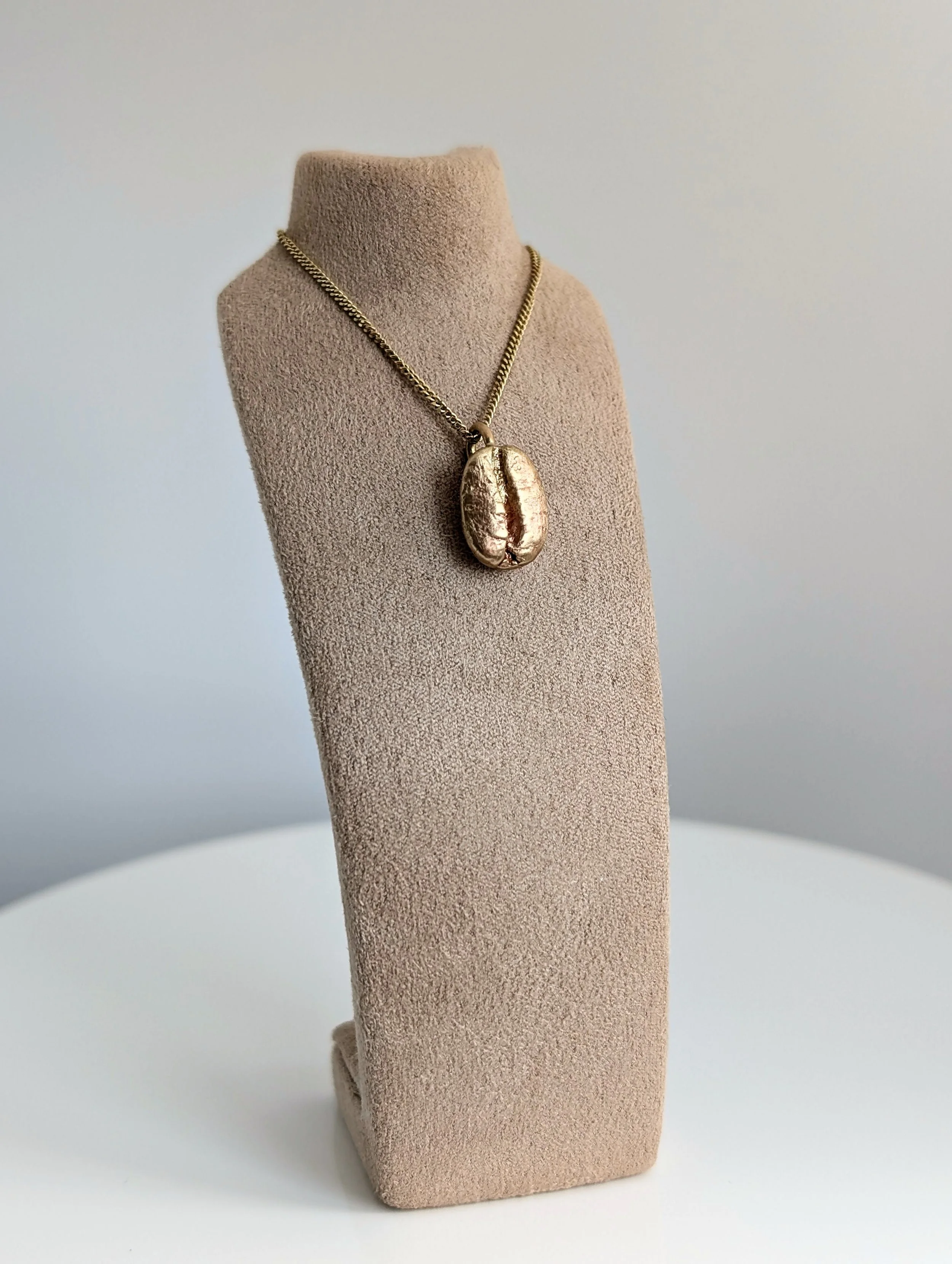 Gold necklace with a pendant shaped like a coffee bean, displayed on a beige jewelry stand.
