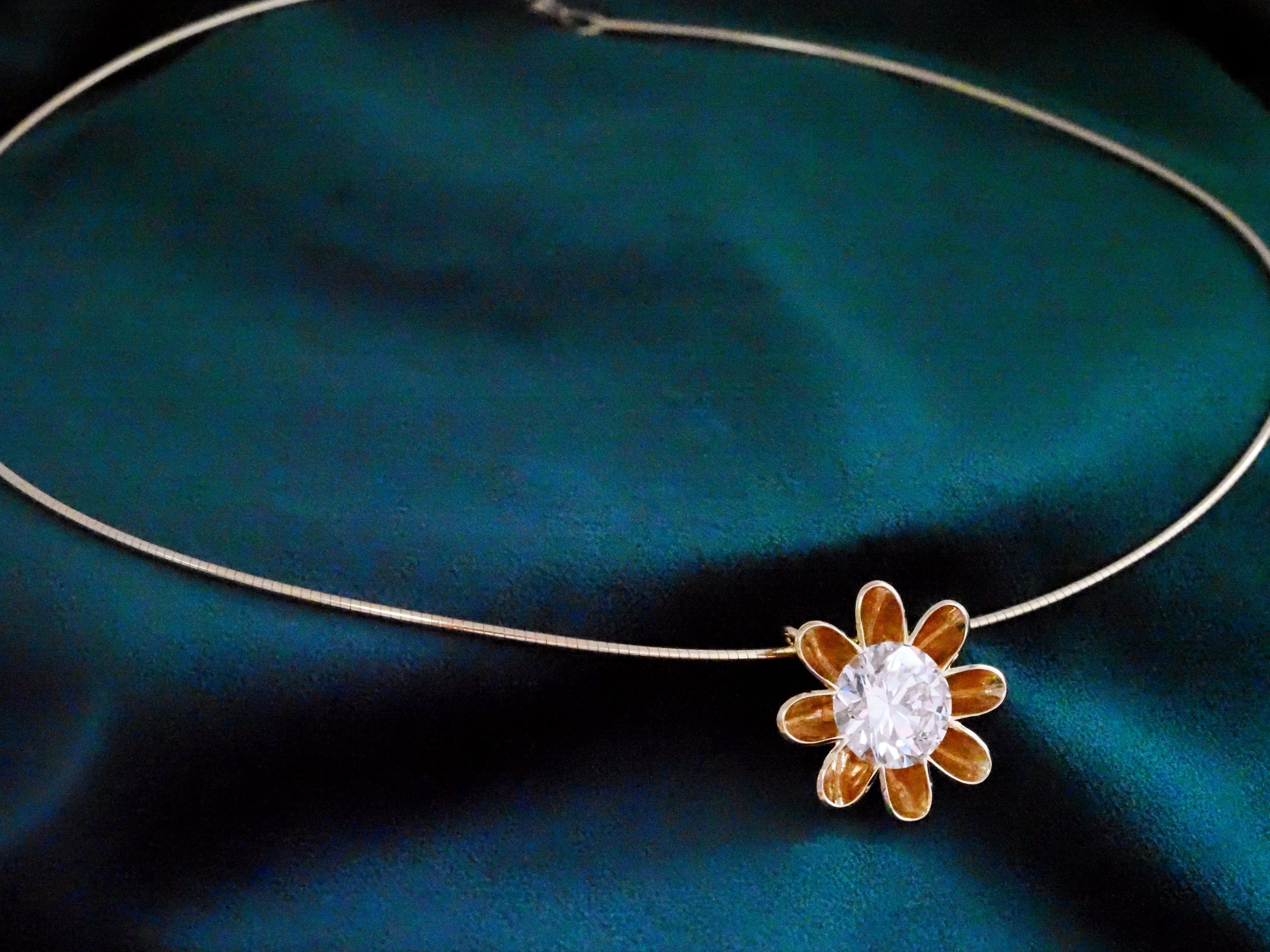 A diamond necklace with a flower-shaped pendant featuring a large central diamond and petal-shaped accents, displayed against a dark teal background.