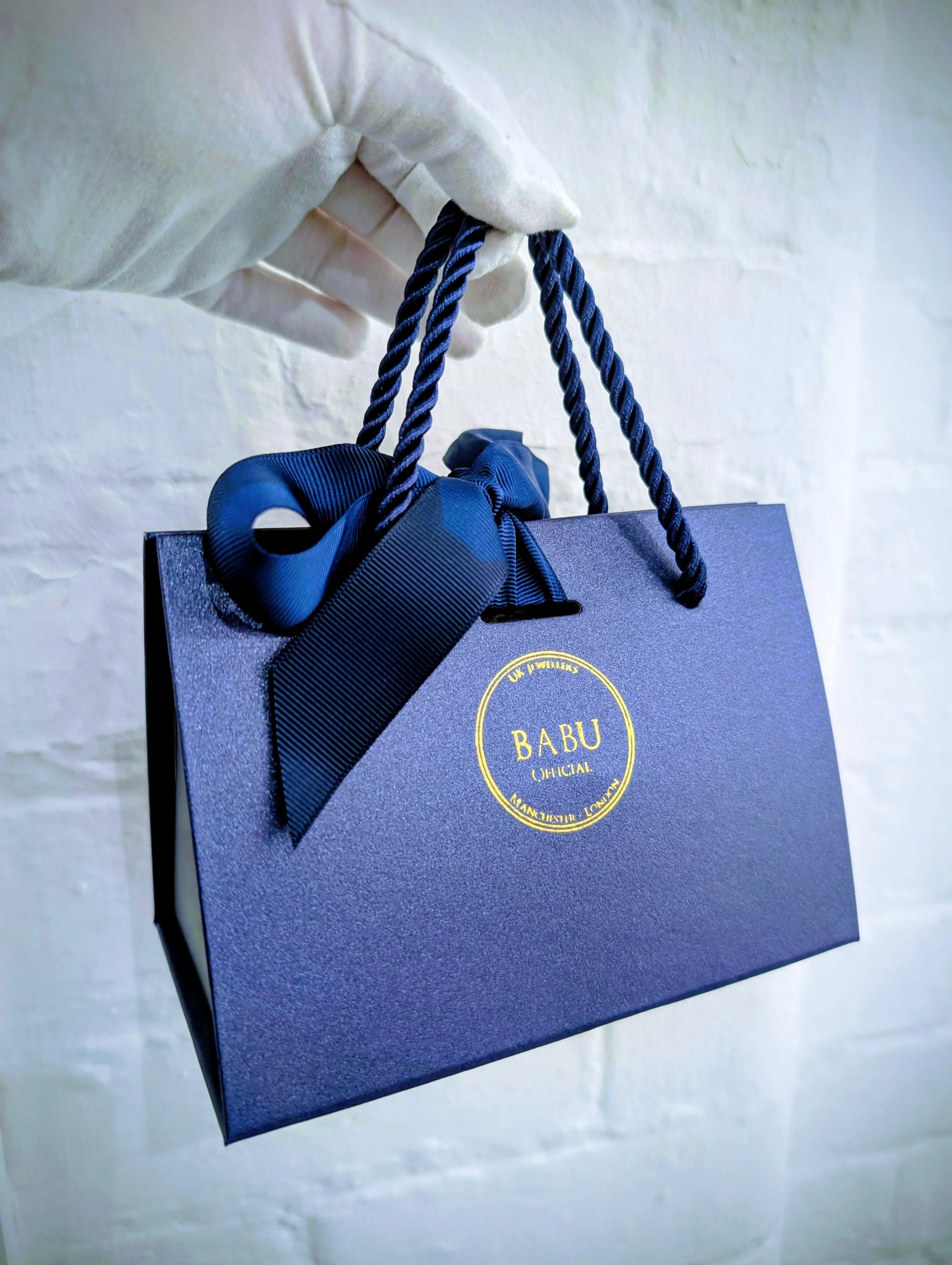 A person wearing a white glove holding a dark blue shopping bag with gold text and a bow.
