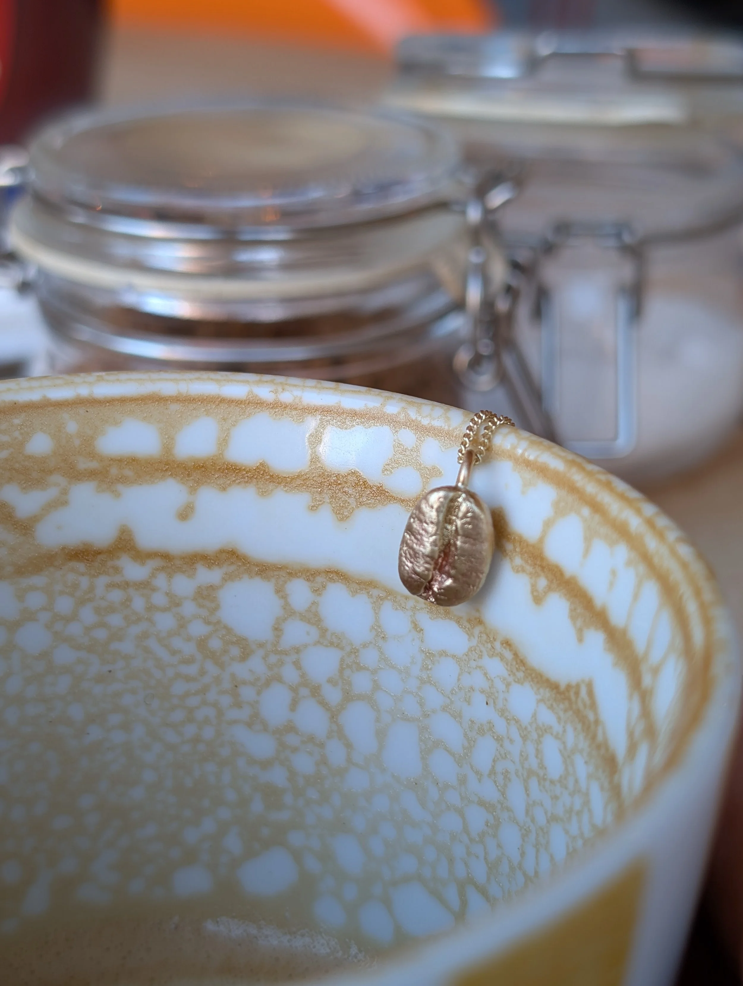 Close-up of a ceramic cup with a necklace featuring a coffee bean pendant hanging over the edge. In the background, there are various glass jars with metal clasps.