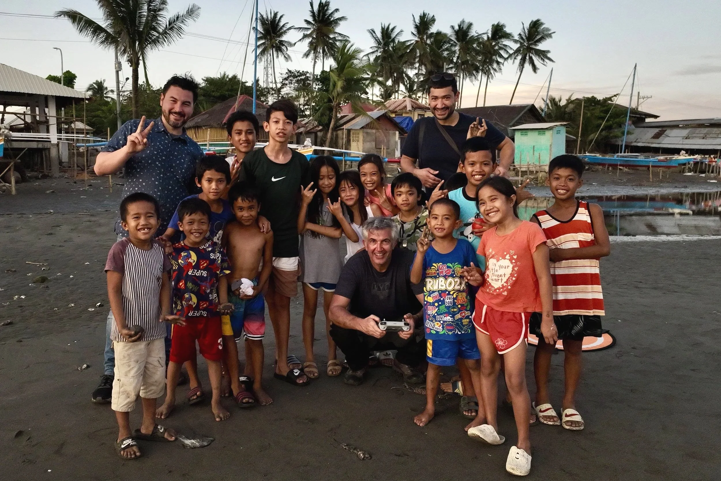 A group of children and two adult men smiling and posing for a photo on a beach with palm trees in the background.