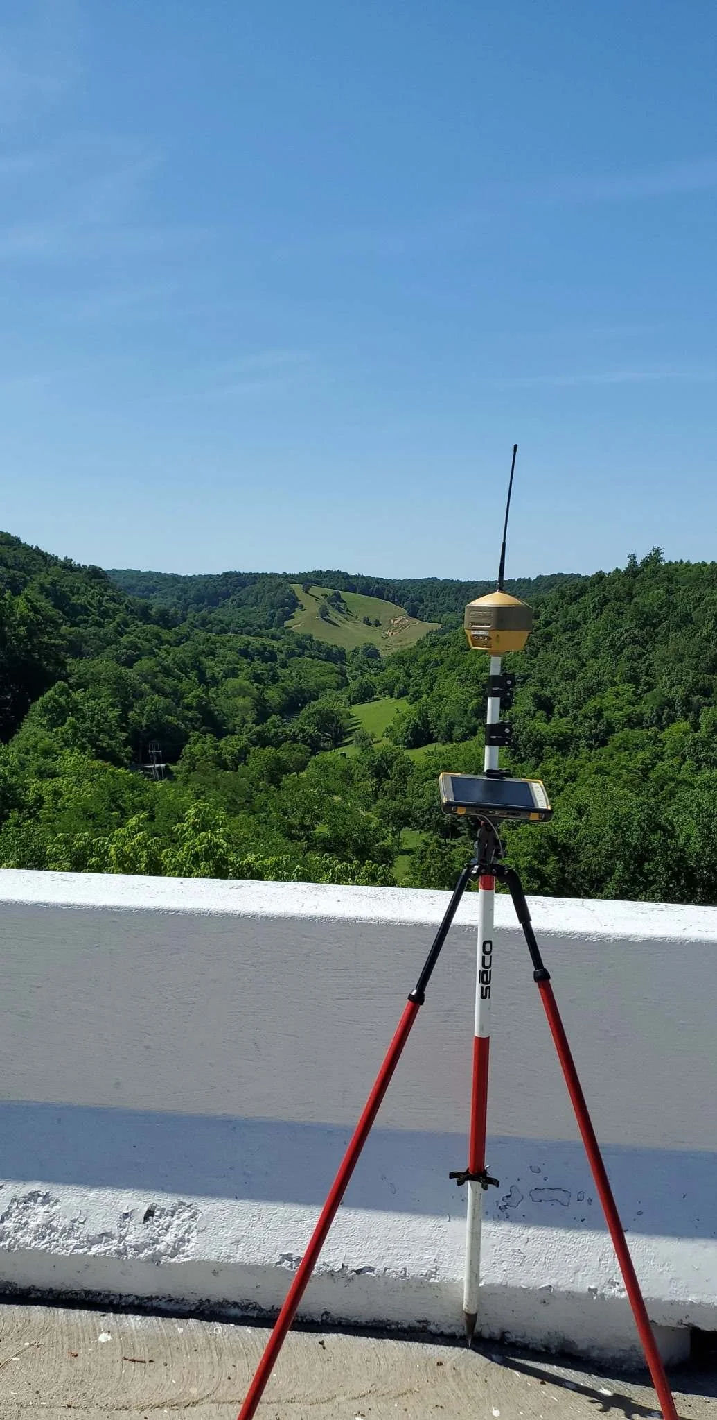 A survey instrument on a tripodal stand with a tablet and antenna, set against a backdrop of green hills and a clear blue sky.