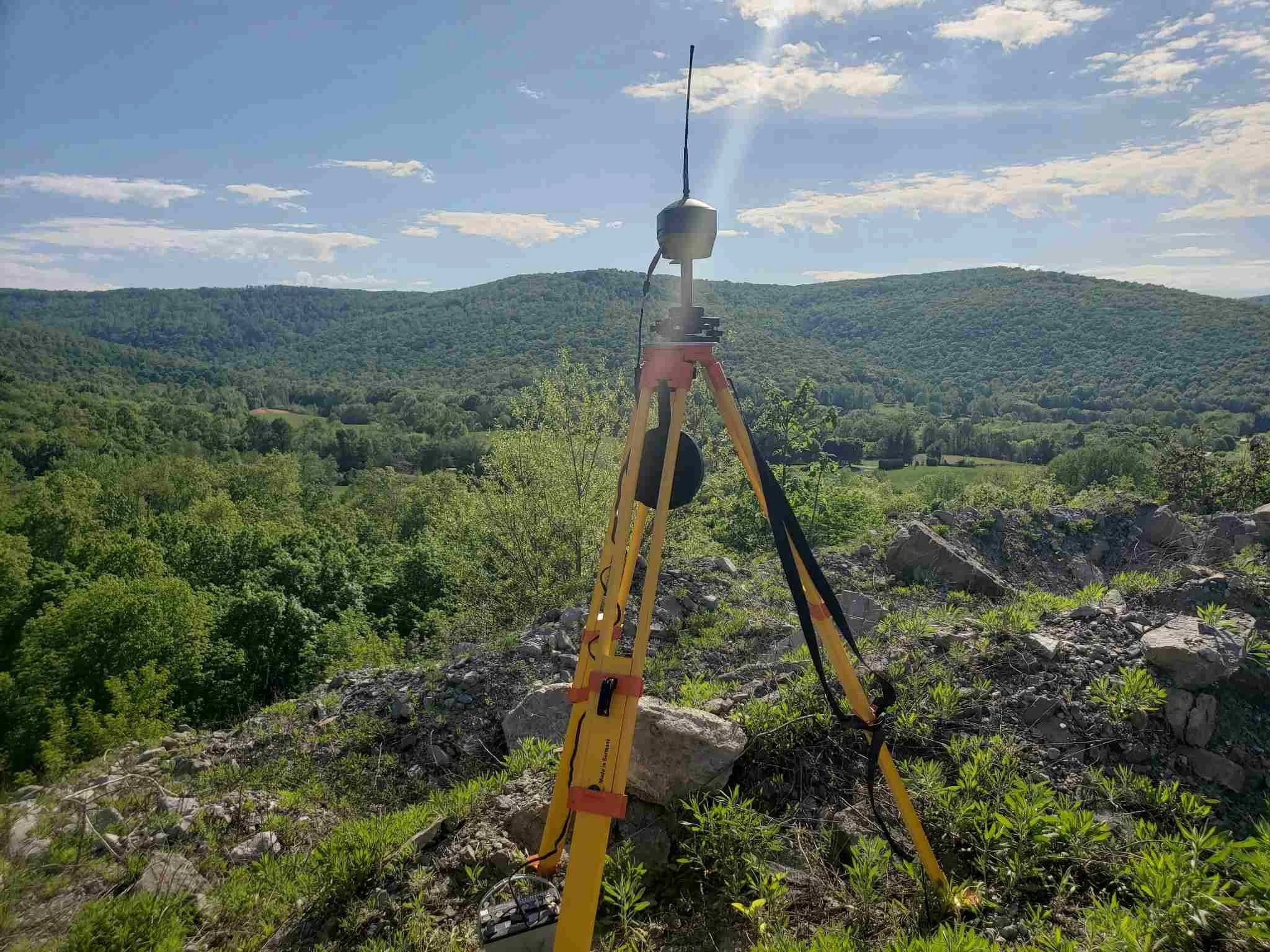 Surveying equipment set up on rocky terrain overlooking a lush green valley with hills under a partly cloudy sky.