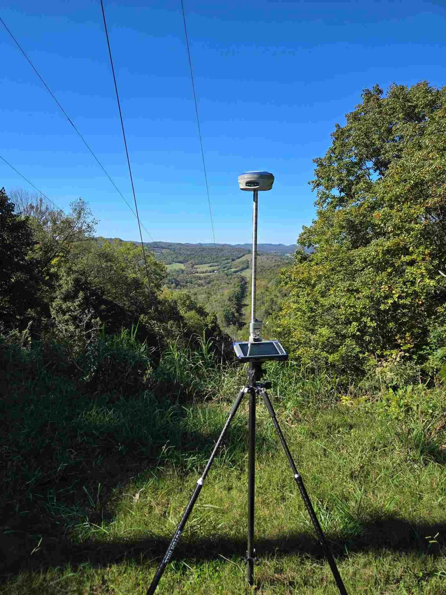 A wildlife tracking camera mounted on a pole and tripod, with a GPS device attached, set in a green outdoor area with trees and mountains in the background.