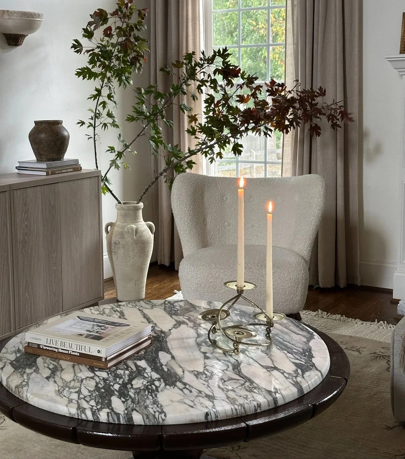 A cozy living room corner featuring a marble-topped round coffee table with books, a candelabrum with lit candles, a white textured armchair, a large vase with a leafy plant, a wooden sideboard, and a window with beige curtains.