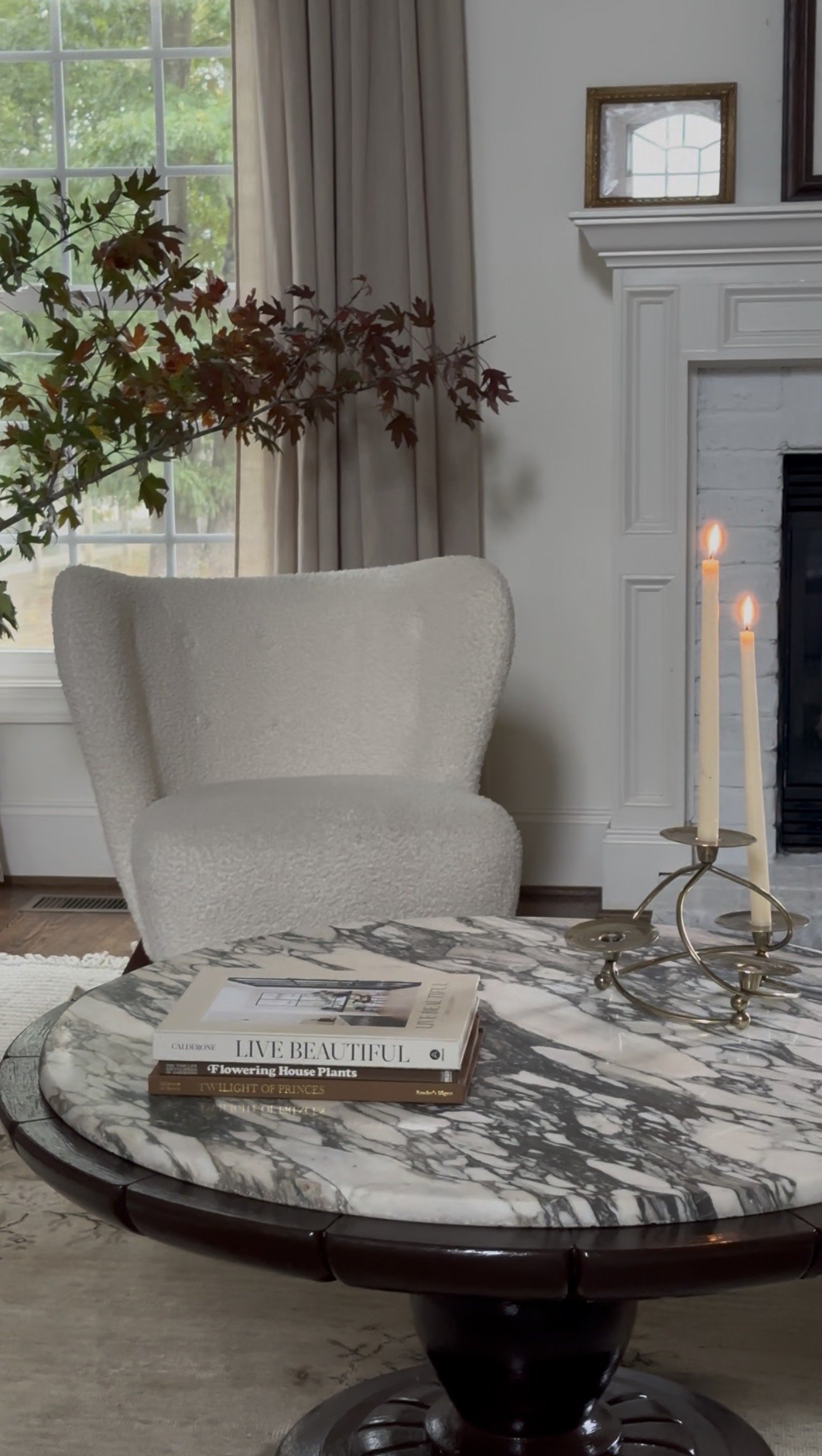 A living room corner with a marble-topped round table, books, a beige armchair, a window with beige curtains, a white brick fireplace, and two lit candles in a candelabrum.