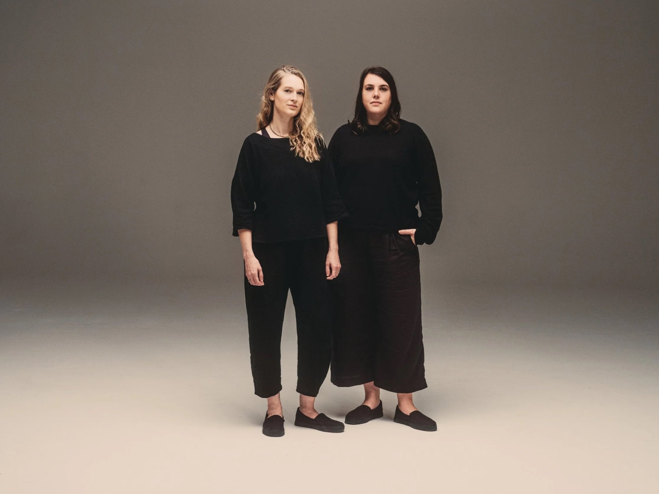 Two women standing against a plain gray background, both dressed in black, with one woman having blonde curly hair and the other having dark curly hair.