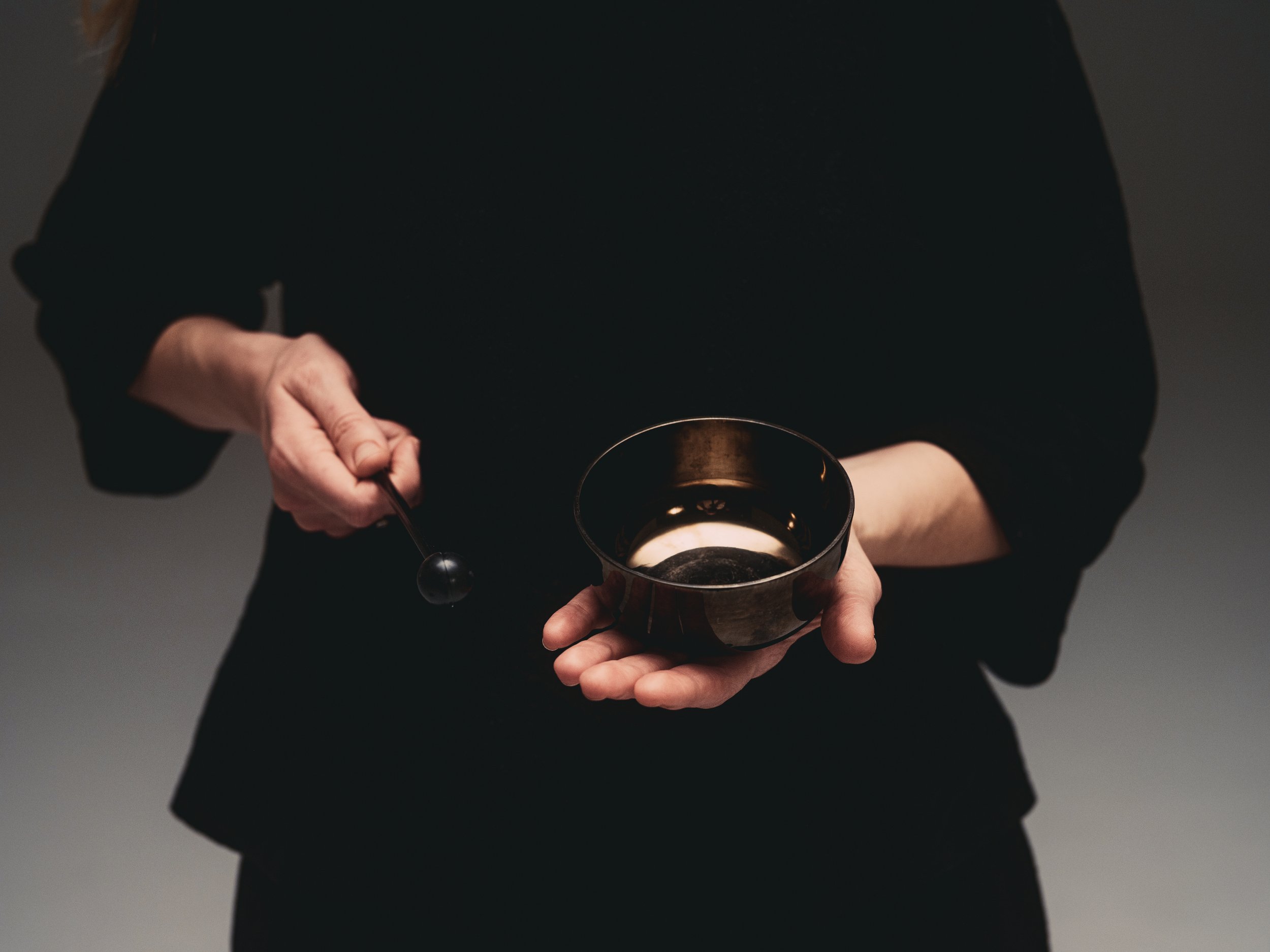 Person in black clothing holding a musical singing bowl and a black beater. The photo is taken mid strike of the bowl.