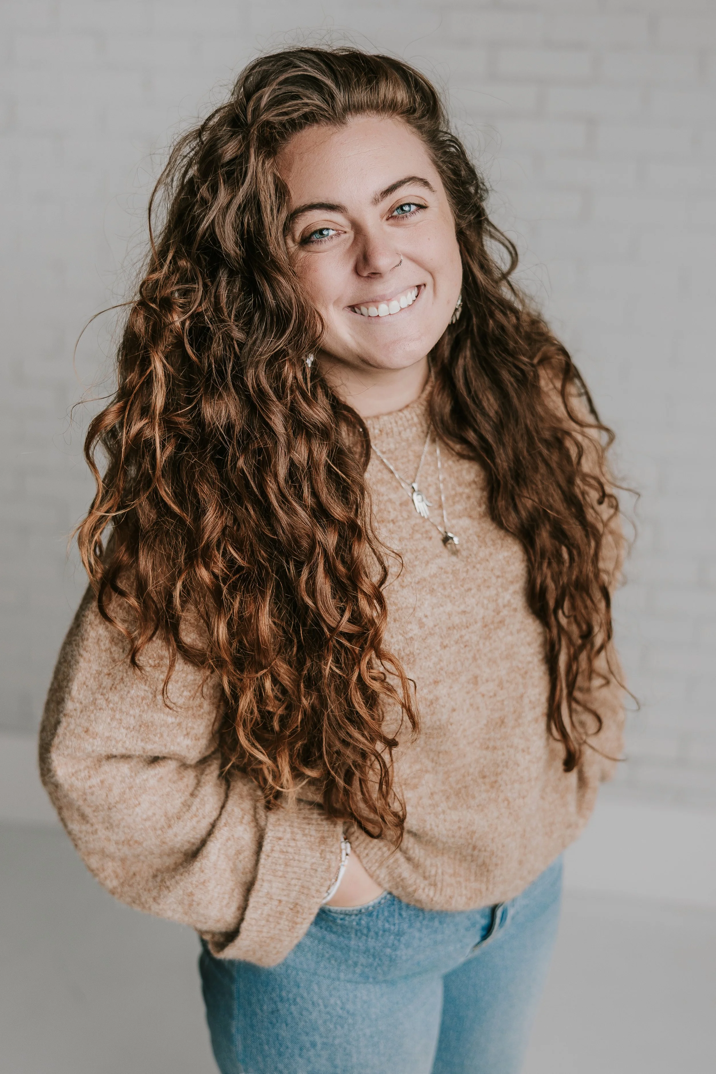 Young woman with long curly hair smiling, wearing a tan sweater and blue jeans, standing in front of a white brick wall.