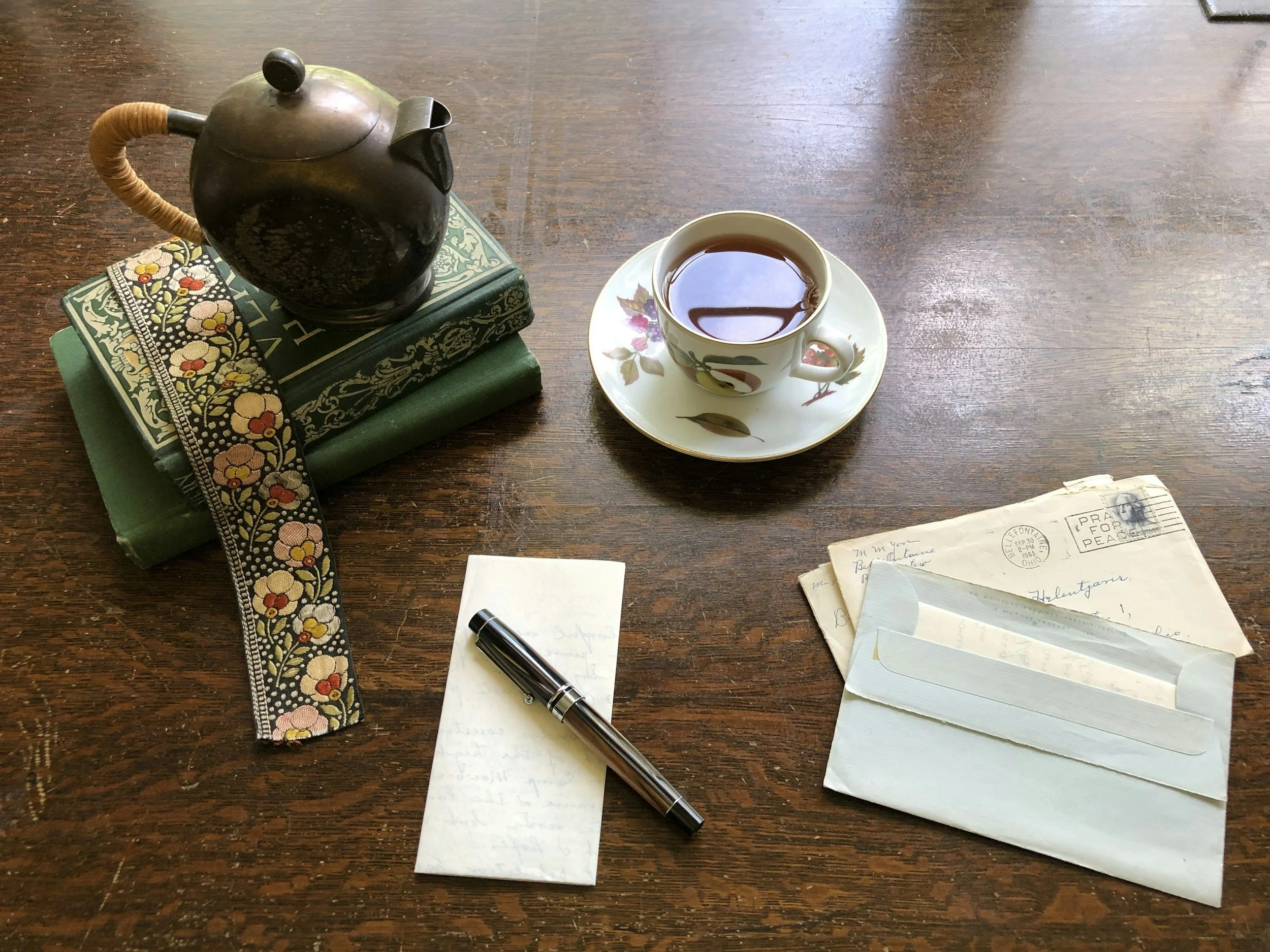 A wooden table with a black teapot on top of two closed books, including a green one and a decorative book with floral print. There is a floral-patterned cup and saucer filled with tea, a small notepad with handwritten notes, a black pen, and two envelopes, one open with a letter inside.