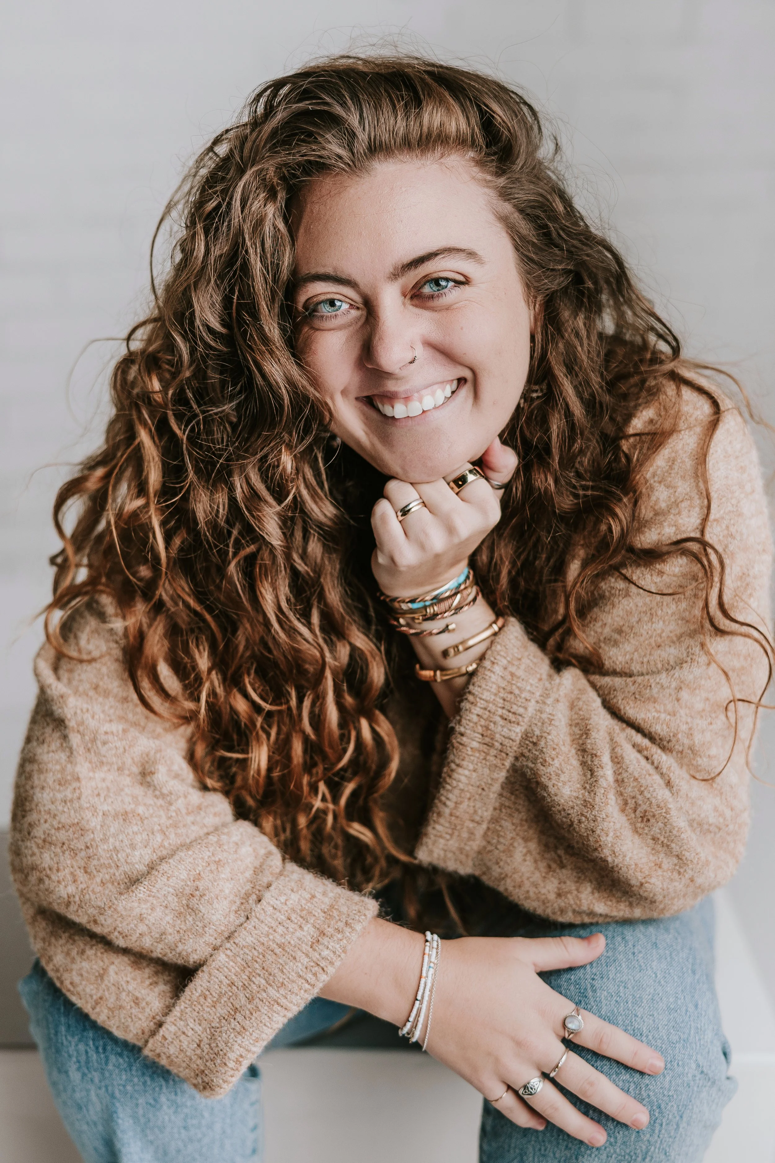 A young woman with long, curly brown hair, blue eyes, and a nose ring, smiling at the camera. She is wearing a tan sweater, multiple bracelets, and rings, and is sitting with her chin resting on her hand.