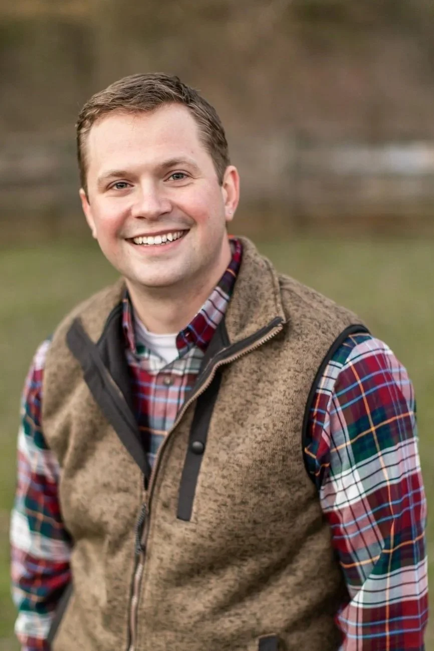 A smiling man wearing a plaid shirt and a brown vest standing outdoors with blurred background.