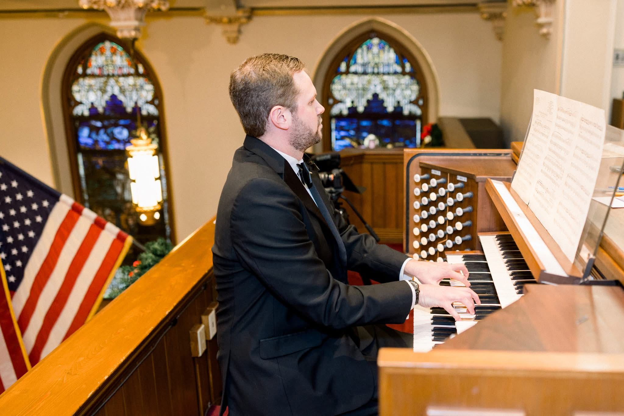 A man in a black tuxedo playing an organ in a church with stained glass windows and an American flag.