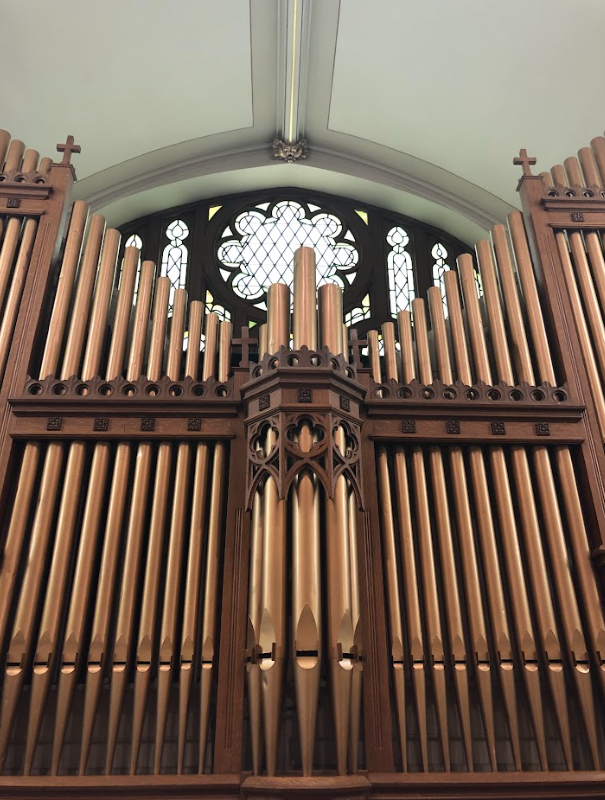 Large wooden church pipe organ with intricate gothic design, situated below a stained glass window with a circular pattern.