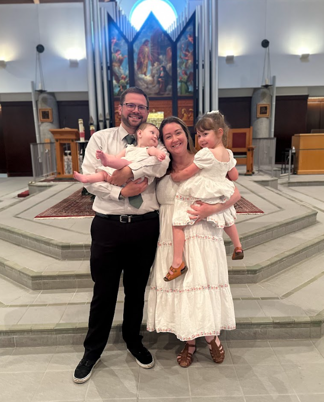 A family of four is standing inside a church, smiling at the camera. The father, wearing glasses and a tie, is holding a baby girl. The mother, dressed in a white dress, is holding a young girl in a light-colored dress. The church features a prominent religious artwork and organ pipes in the background.