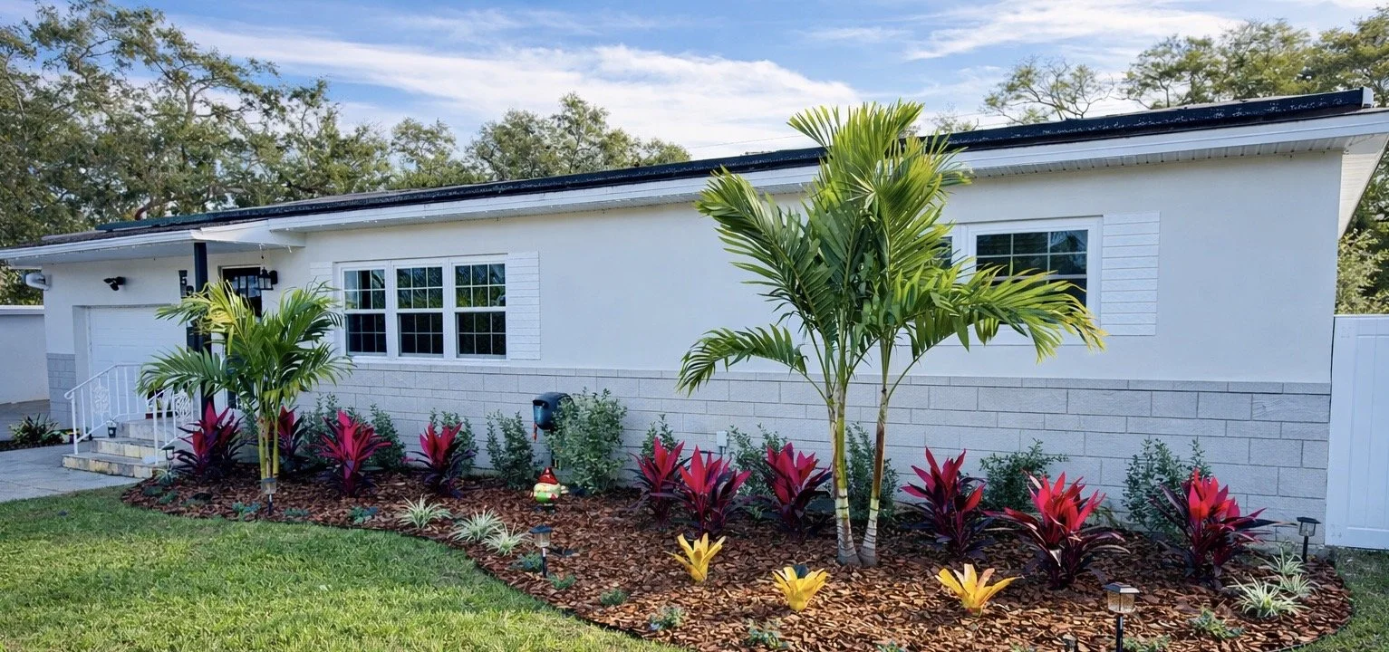 A white house with a garden featuring tropical plants, mulch, decorative lighting, and a manicured lawn.