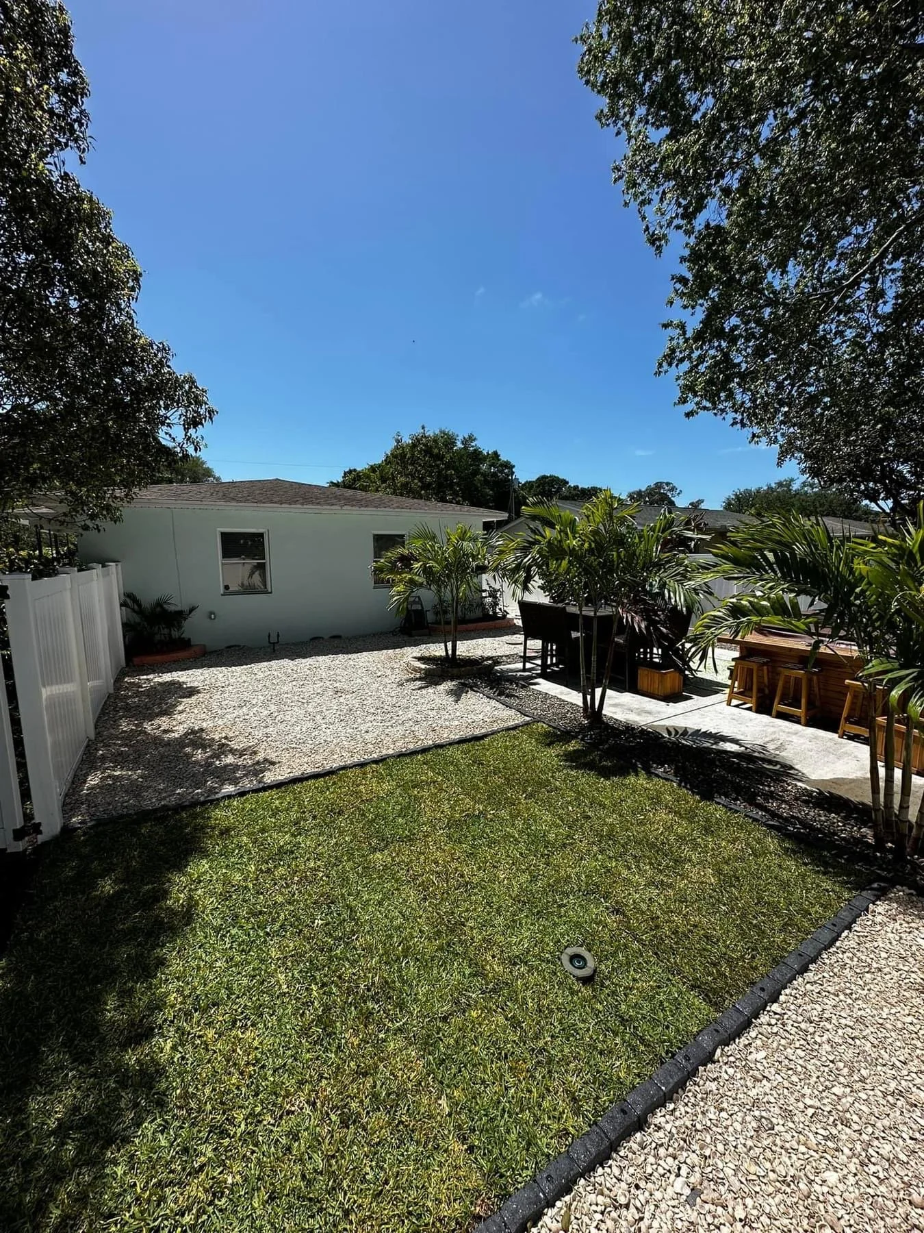A backyard with green grass, palm trees, a gravel area, outdoor furniture, and a white house under a blue sky.