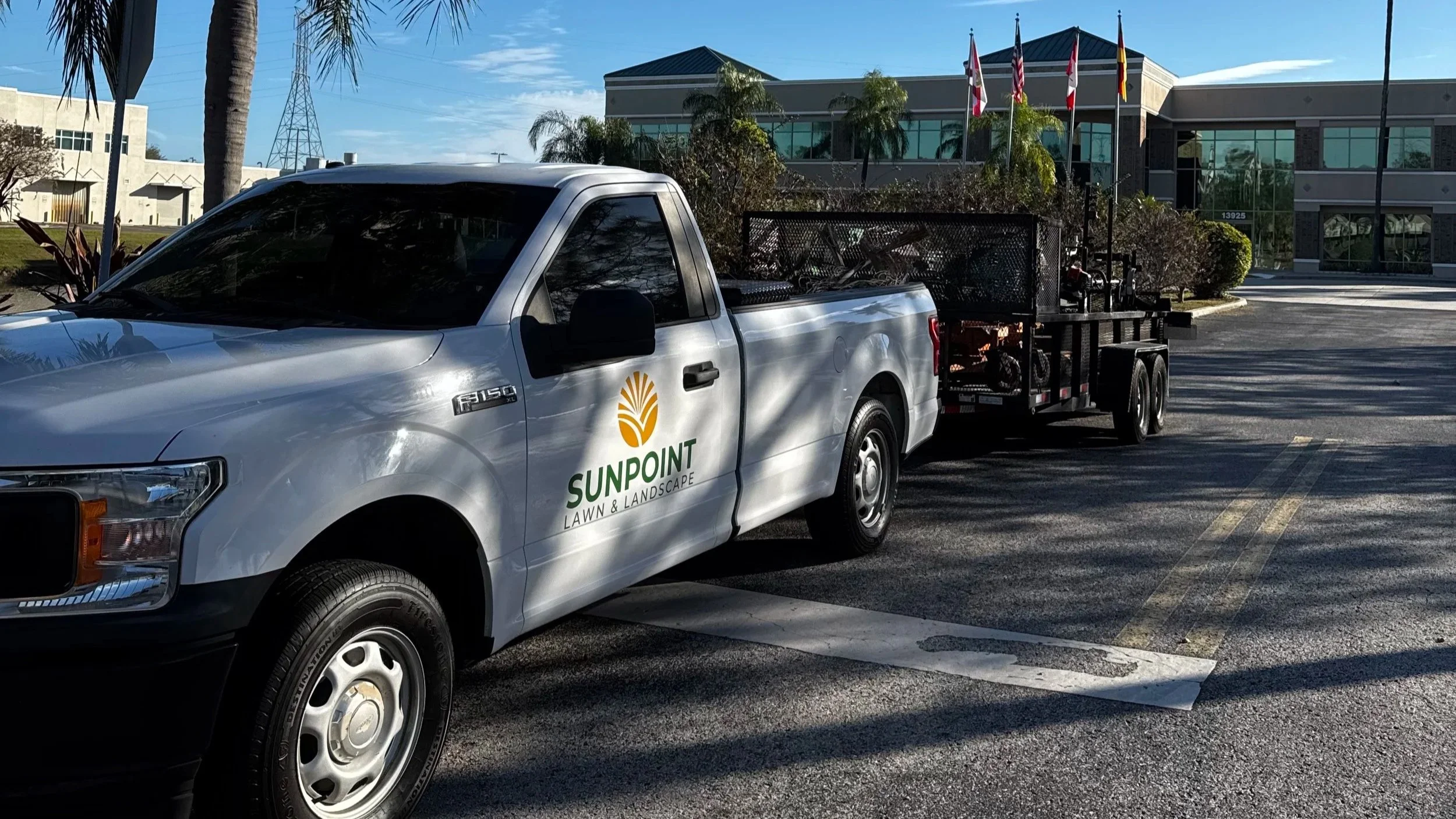 A white pickup truck with 'Sunpoint Lawn & Landscape' logo parked in a parking lot, towing a flatbed trailer filled with landscaping equipment.