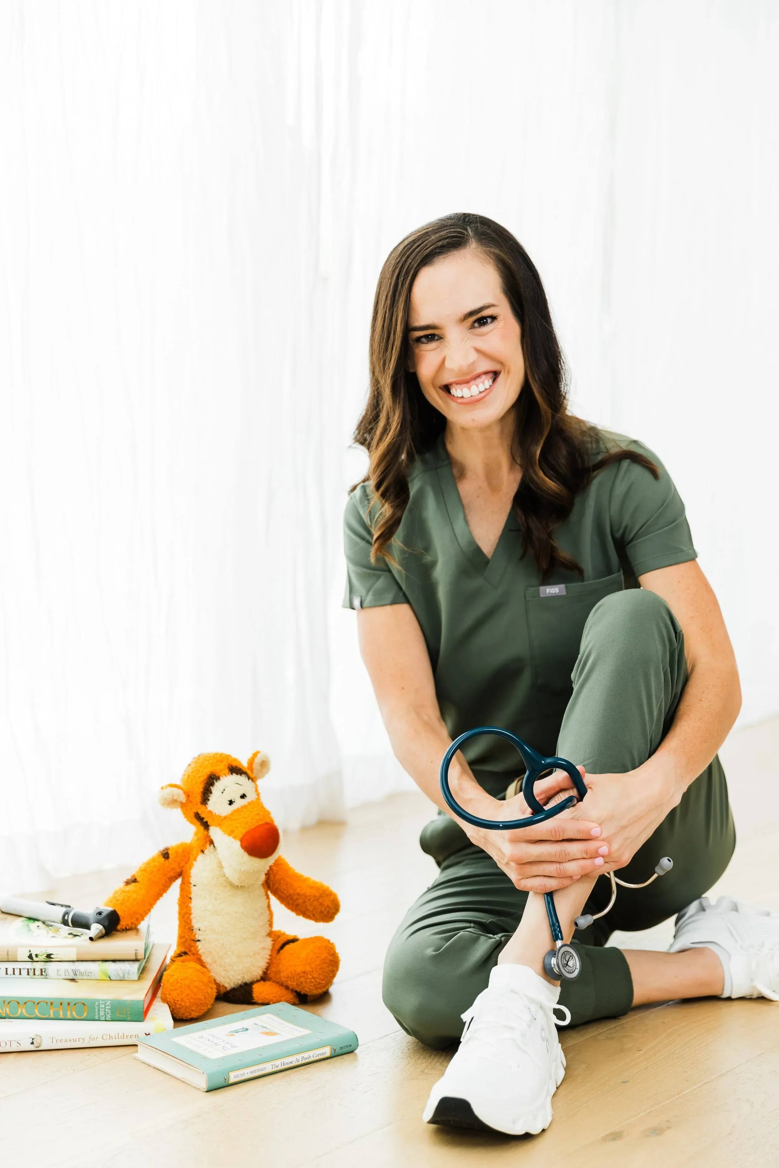 Dr. Jenna Penland in green scrubs sitting on the floor, holding a stethoscope around her foot and smiling. There are children's books and a plush toy on the floor beside her.