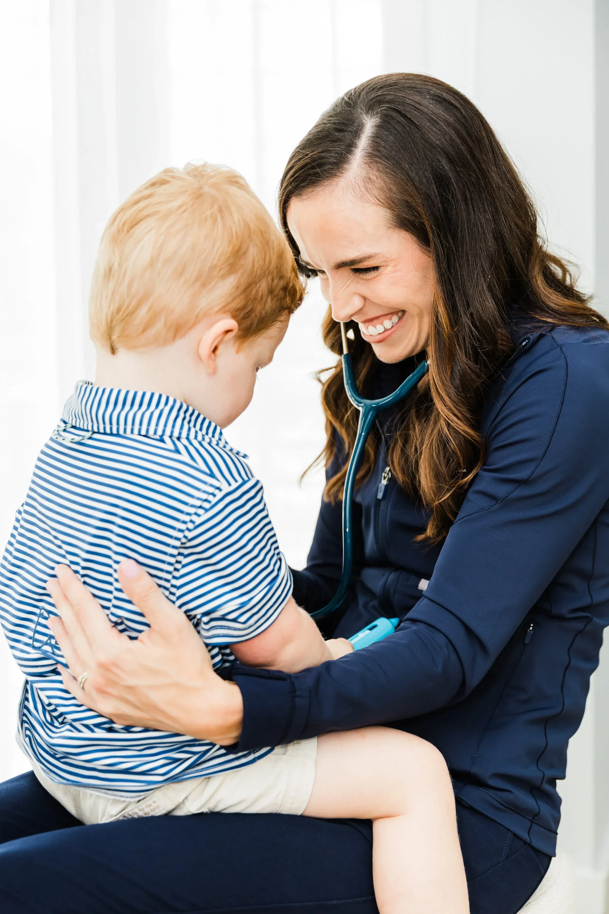 Dr. Jenna Penland with a stethoscope around her neck, smiling and holding a young boy who is sitting on her lap, in a medical office setting.