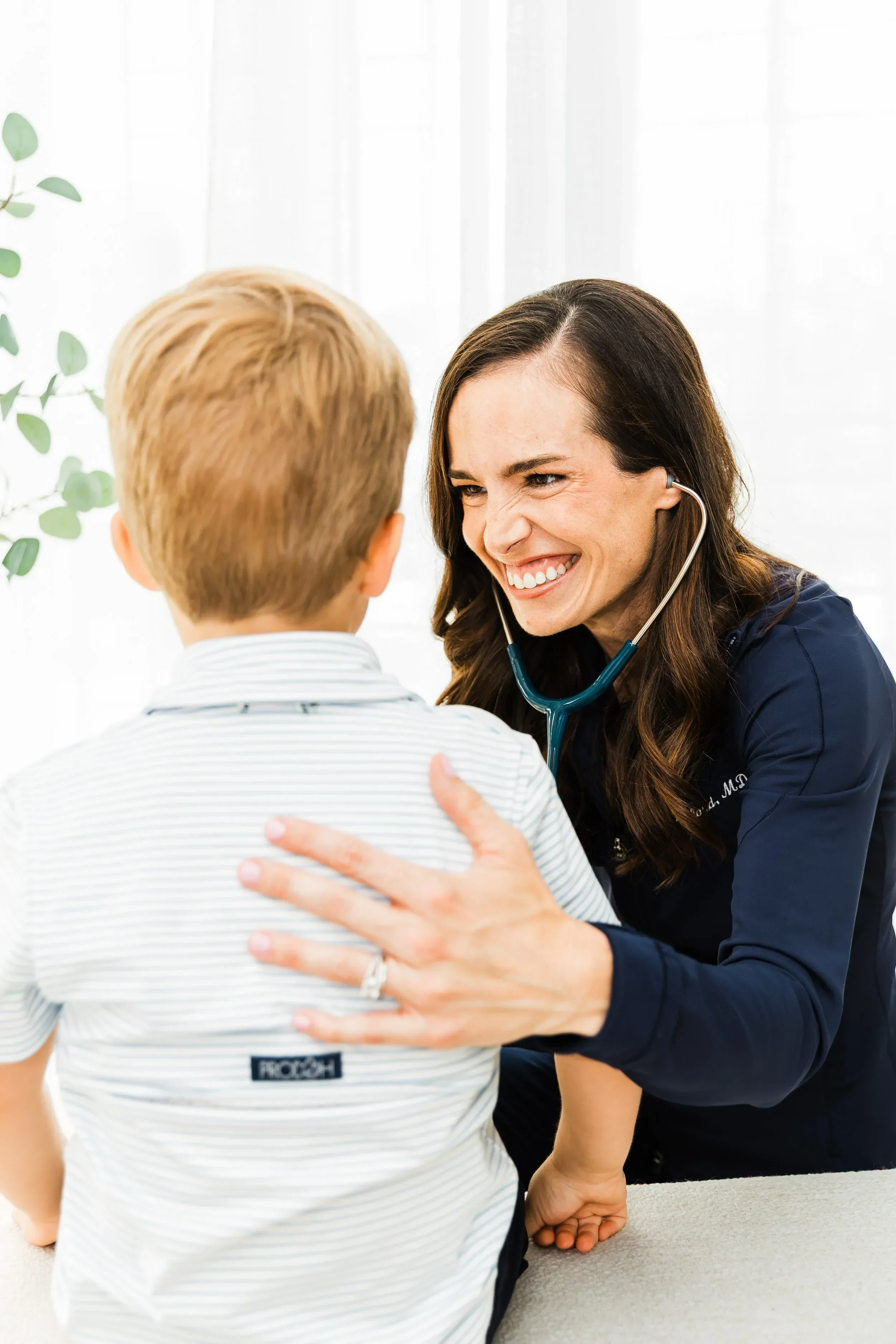 Dr. Jenna Penland smiling and talking to a young boy during a checkup, with her hand on his shoulder and a stethoscope around her neck.