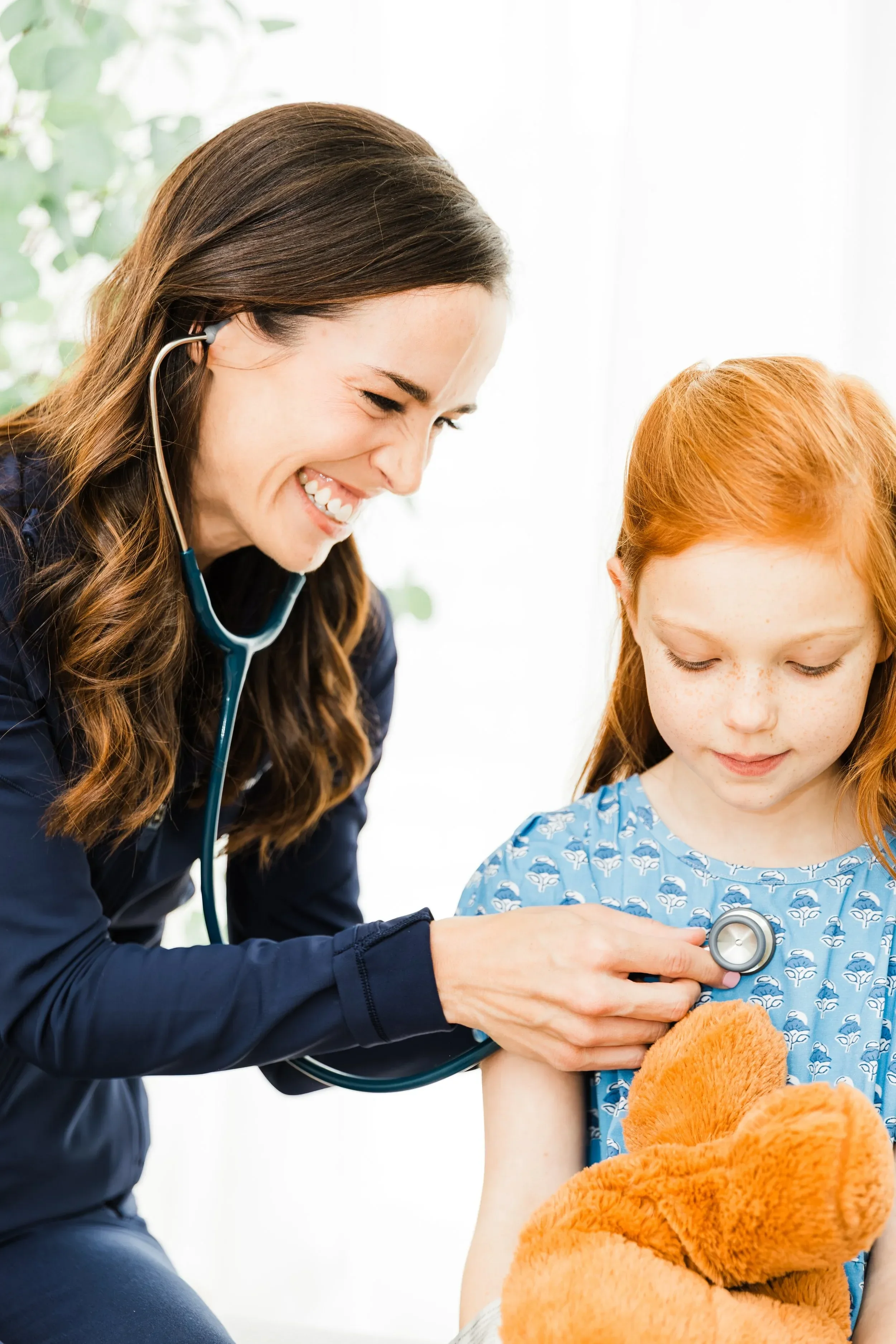 Dr. Jenna Penland using a stethoscope to examine a young girl with orange hair and freckles, sitting in a medical setting with bright natural light.