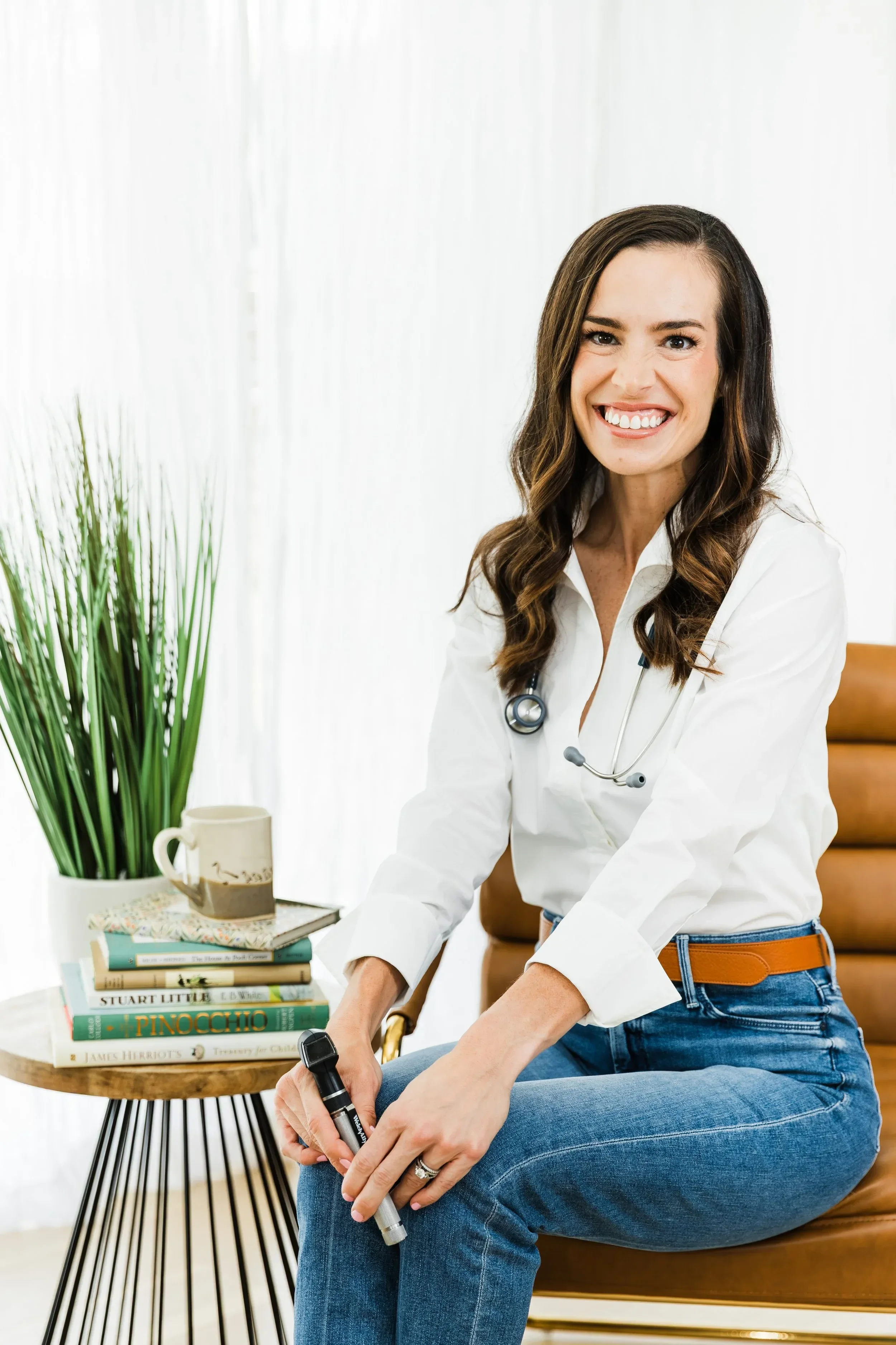 Dr. Jenna Penland in white coat with stethoscope, sitting on a chair, holding a marker, in a bright office with books, mug, and a plant.