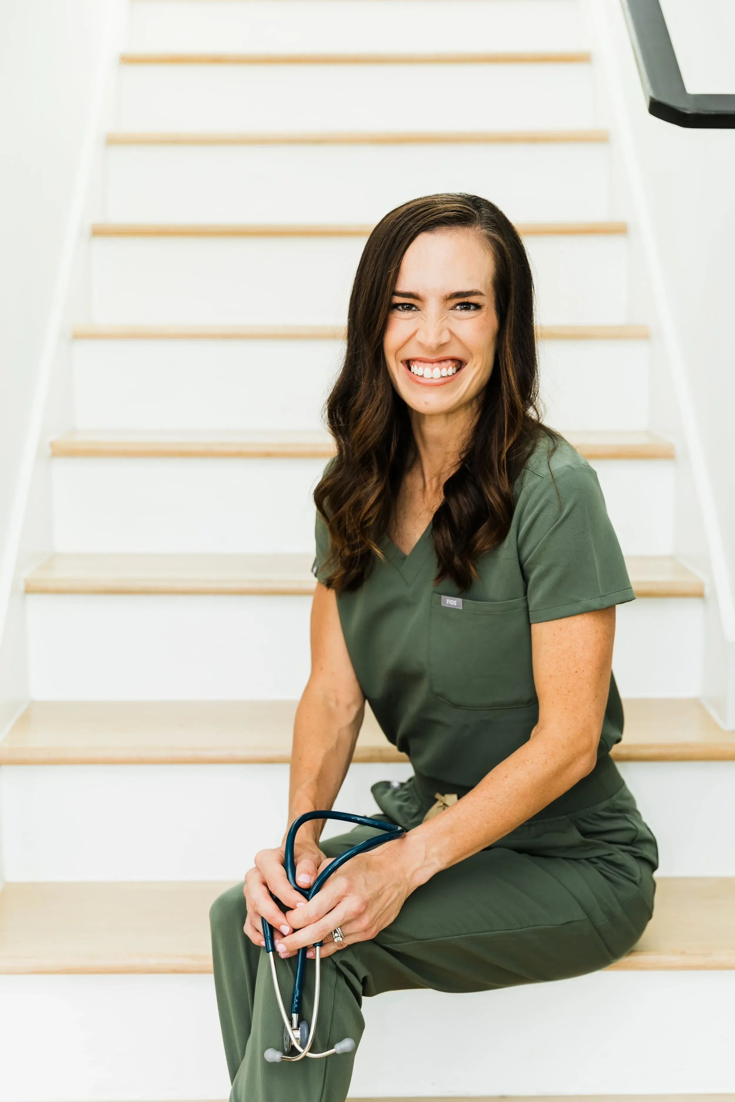 Dr. Jenna Penland in green medical scrubs sitting on a staircase, holding a stethoscope, smiling at the camera.