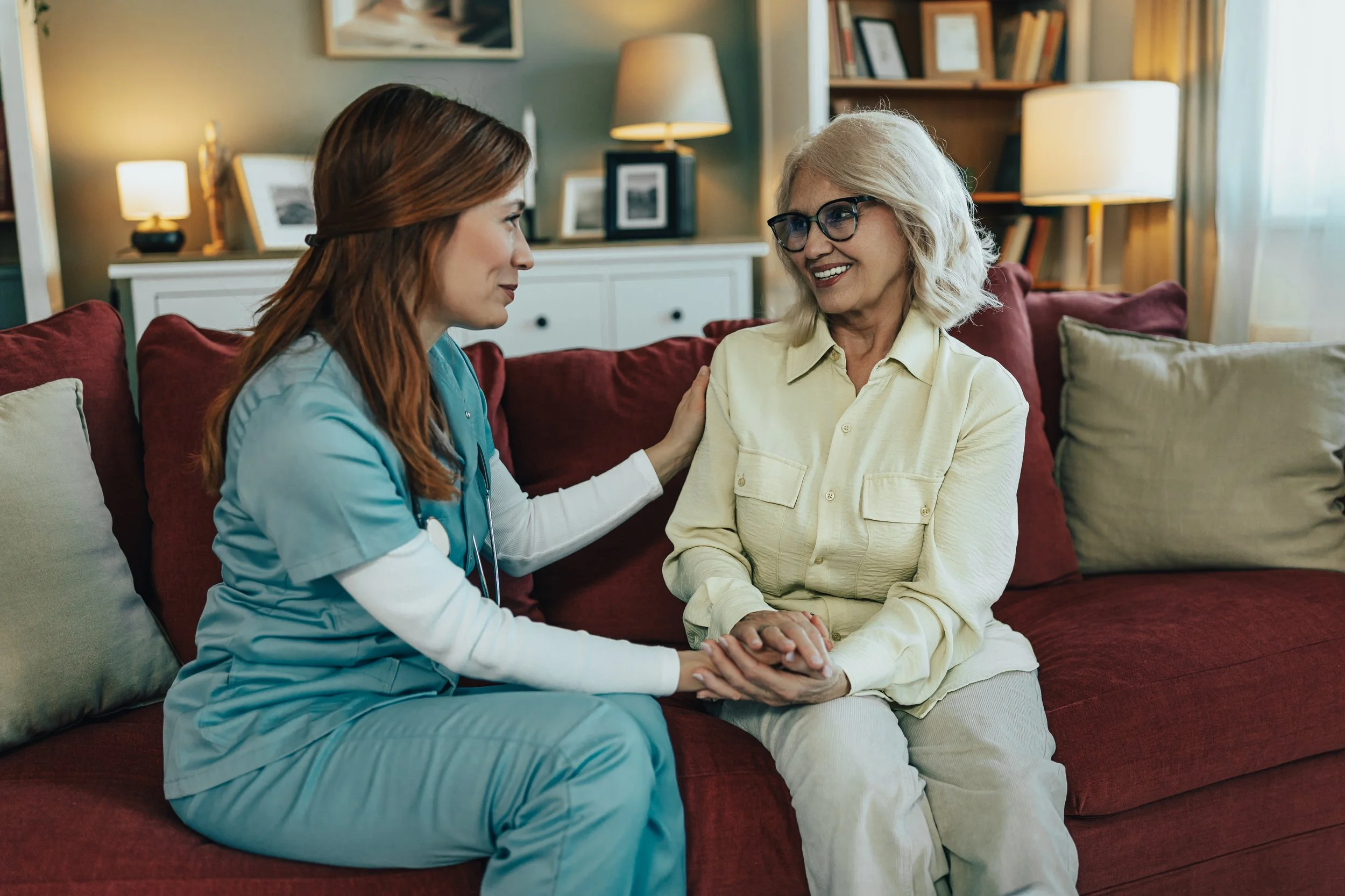 A young female nurse in scrubs shows an elderly woman in a cream-colored outfit something on her hand while sitting on a red couch in a cozy living room.