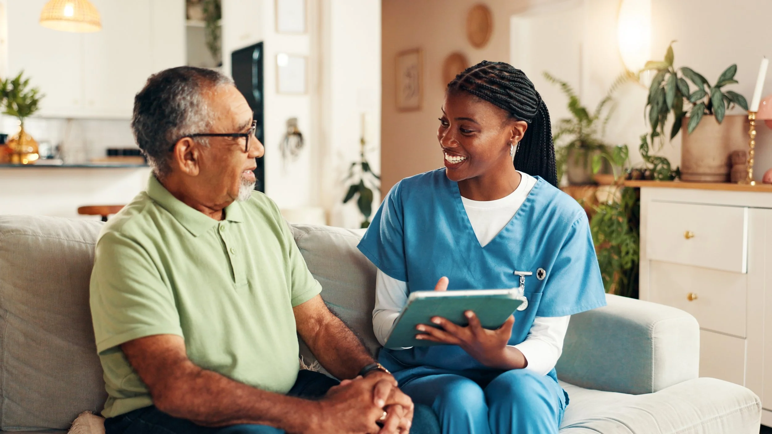 A young female nurse in blue scrubs talking to an elderly man on a beige couch in a living room.