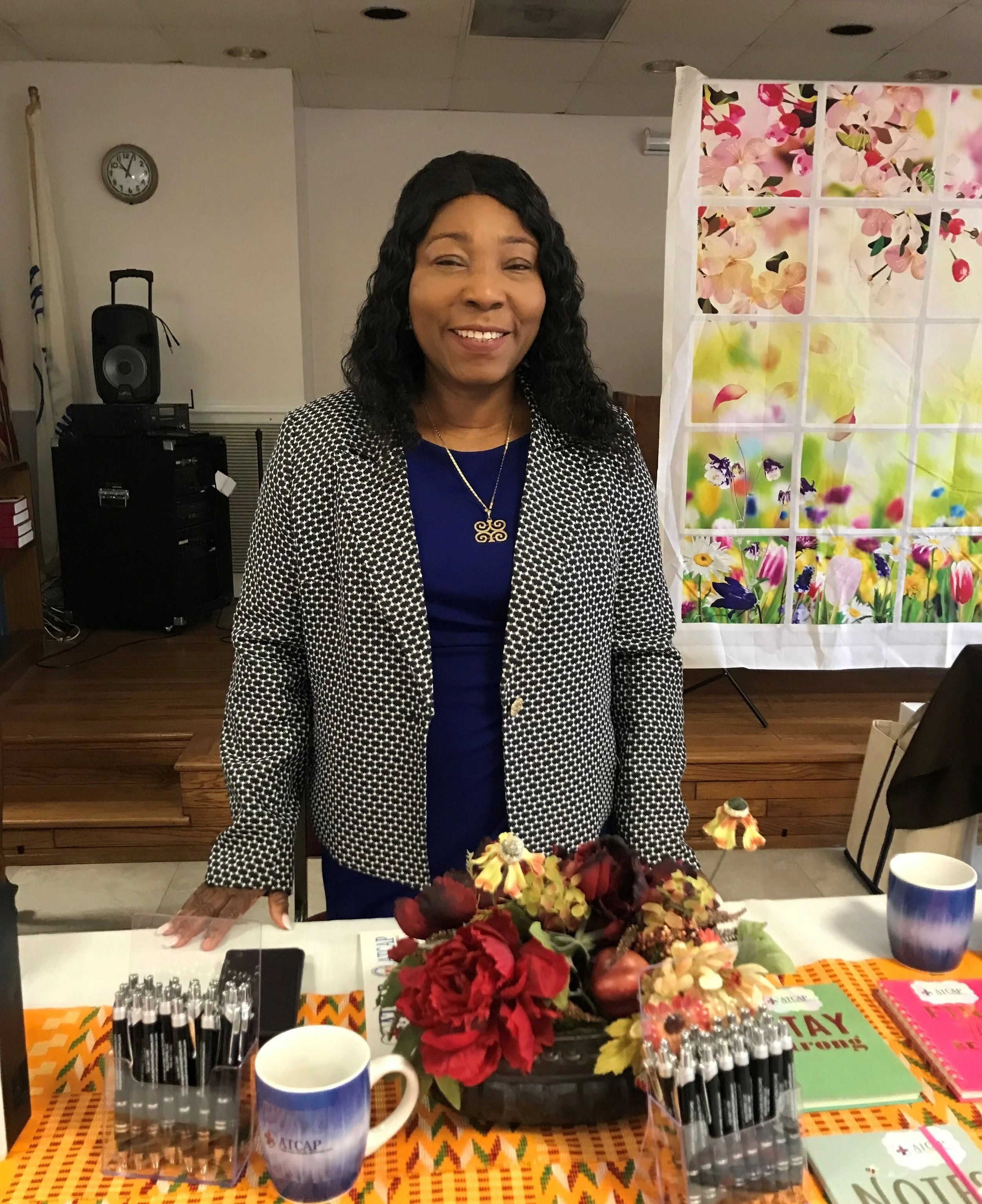 A woman with black wavy hair smiling and standing behind a table decorated with a floral arrangement, pens, notebooks, and cups, in a room with a floral backdrop and a clock on the wall.