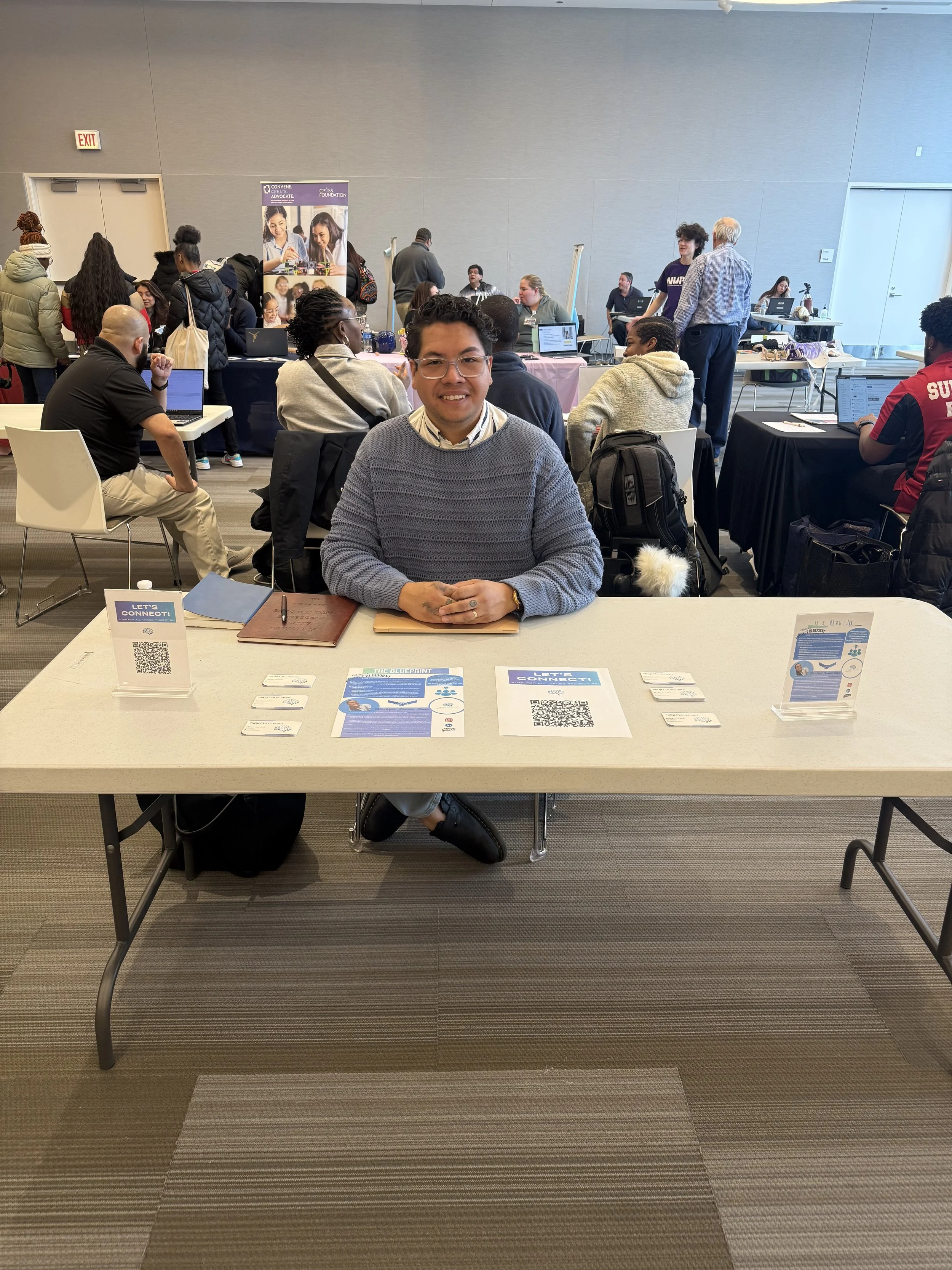 A man sitting behind a table with informational signs and QR codes, at a busy conference or expo, smiling at the camera.