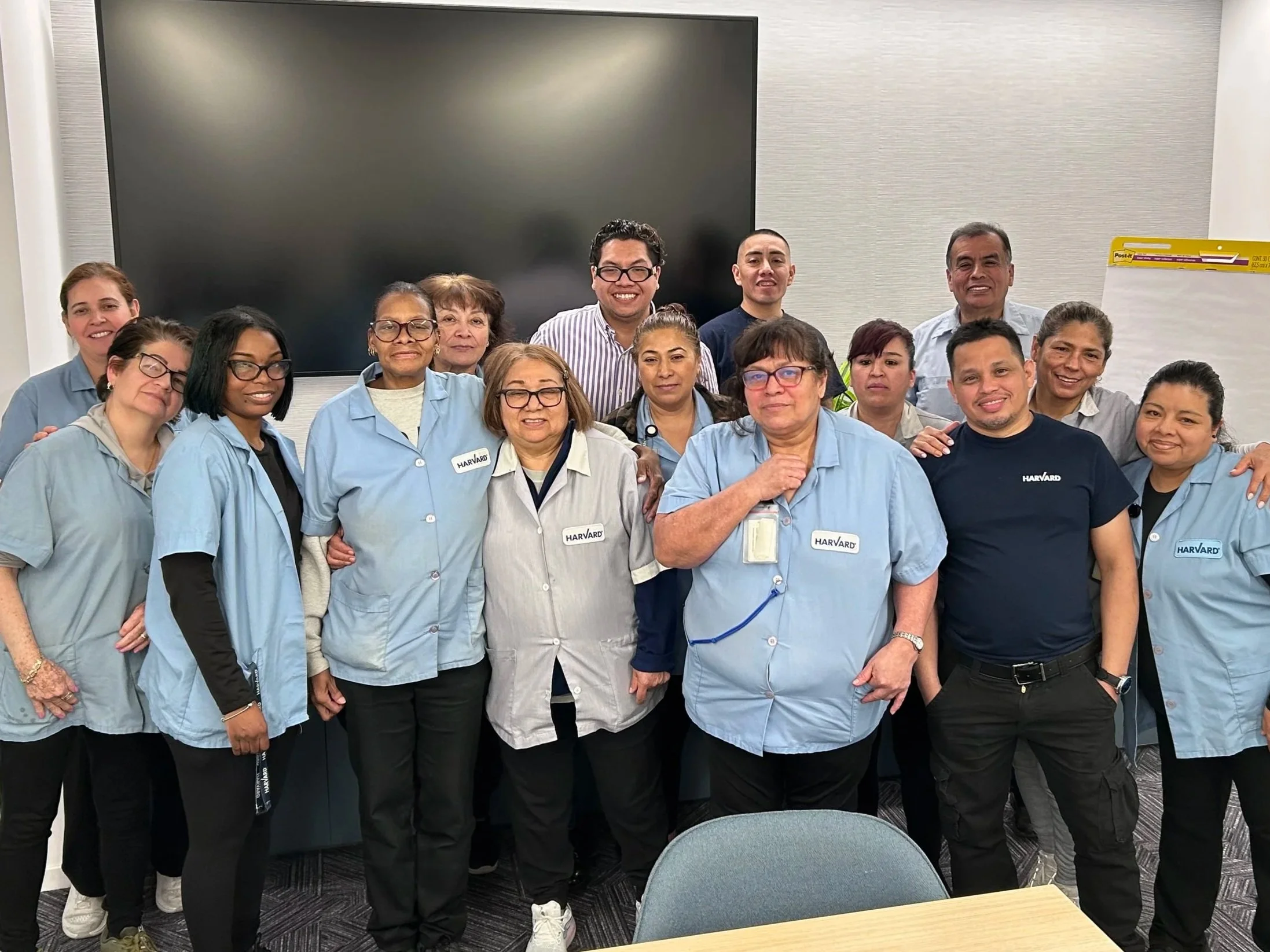 Group of healthcare professionals, including nurses and staff, gathered in a meeting room for a group photo, some wearing blue scrubs with 'Harvard' name tags, smiling and standing in front of a large black screen.