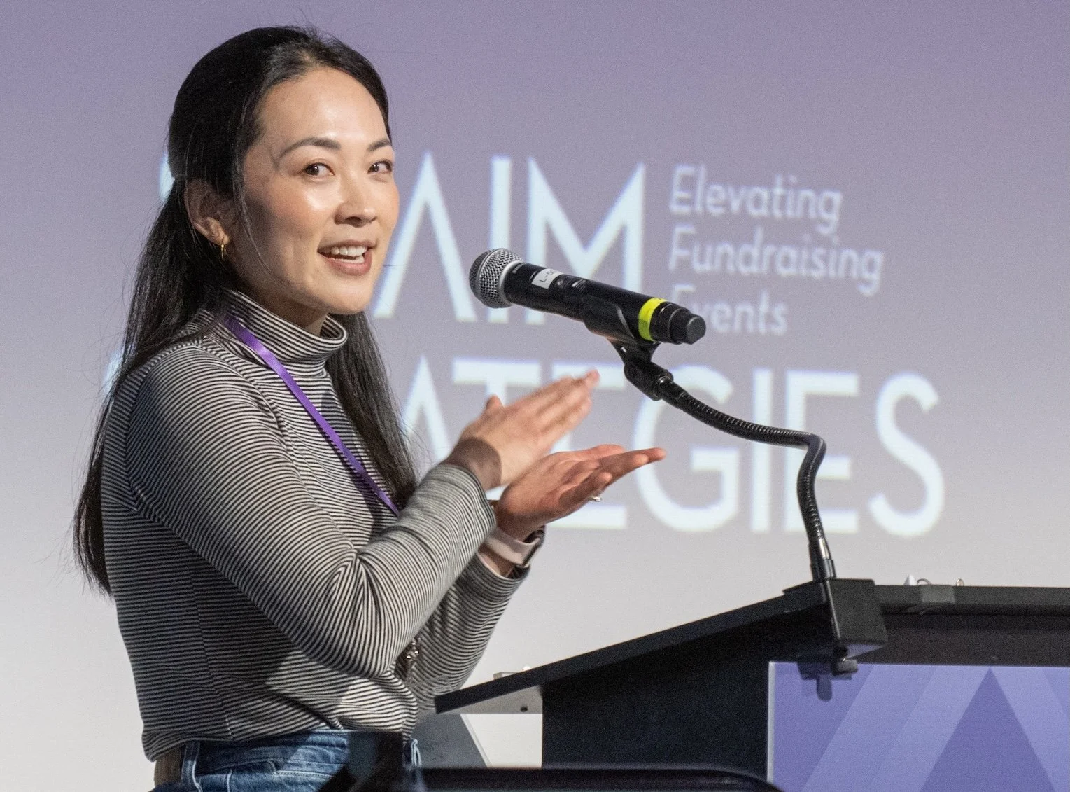Veronica Kim of The Cause Catalyst speaking at a podium with a microphone, in front of a screen with the words 'Aim Elevating Fundraising Clients' and 'Strategies'