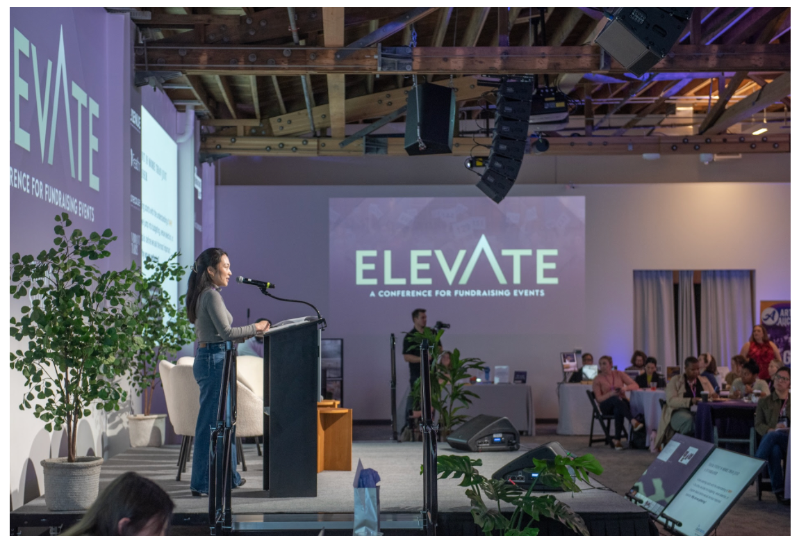 A woman speaking at a conference stage with a microphone, in front of a large screen displaying the event name 'ELEVATE, A Conference for Fundraising Events.' The audience sits at tables, and a cameraman records the event. There are potted plants on stage and electronic screens displaying information.