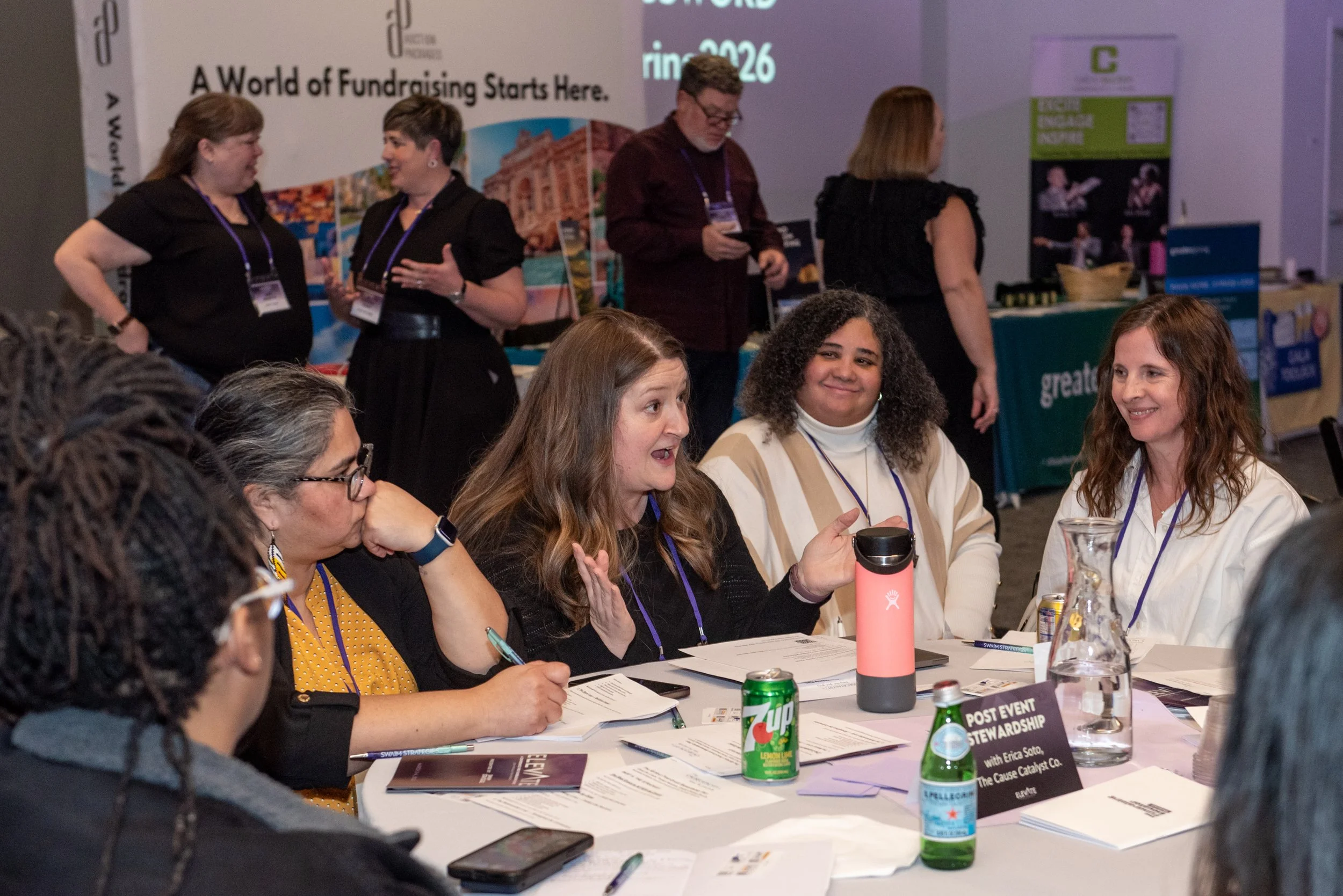 Group of women sitting around a table engaging in a discussion at a conference, with posters and booths in the background.