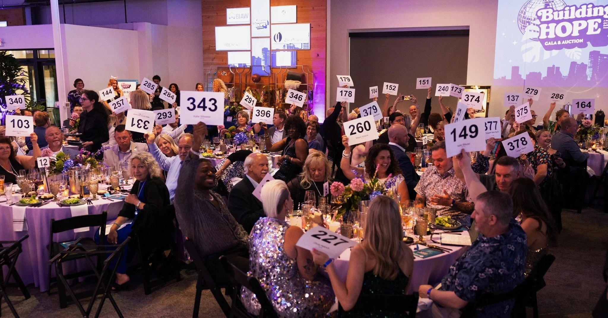 People at a fundraising event holding numbered signs in a banquet hall with round tables and colorful floral centerpieces, and a large screen on the wall displaying 'Building Hope Gala & Auction'