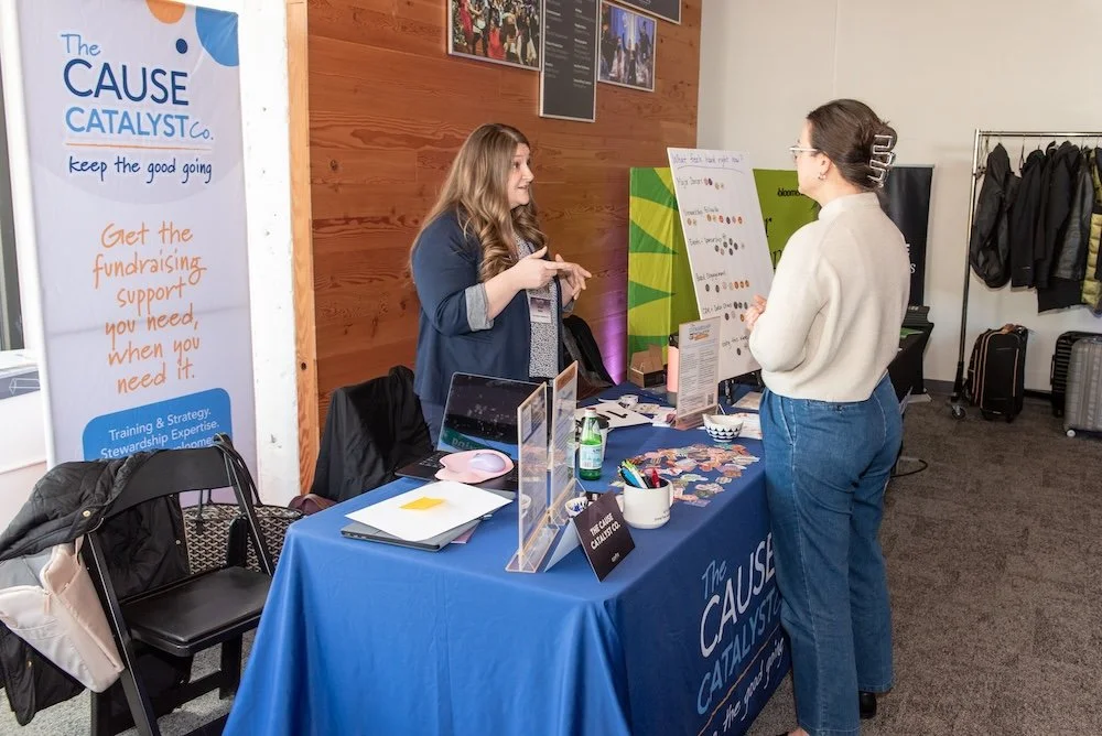 Two women engaged in conversation at a booth promoting The Cause Catalyst, with a blue tablecloth, informational materials, and a display board, in an indoor setting with wooden paneled wall and coat rack.