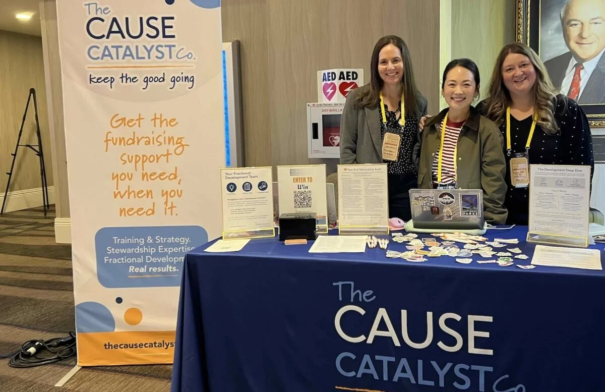 Three women standing behind a table with informational materials at a fundraiser event for The Cause Catalyst. The table has a blue tablecloth with the organization's name, and a banner stands nearby with details about fundraising support and training.