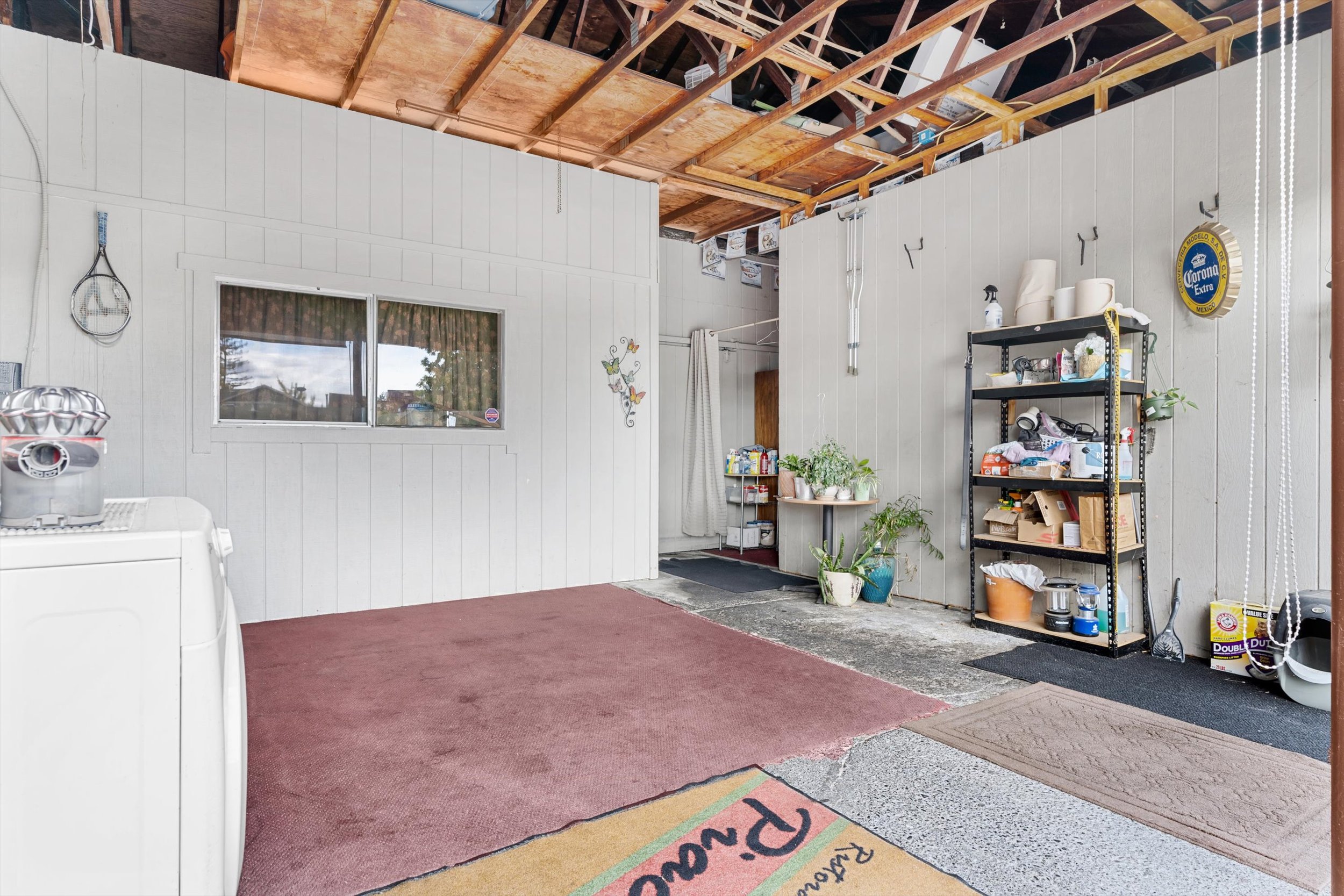 An unfinished garage with white wooden walls, a partially exposed wooden ceiling, a window, a black metal shelf filled with various items, potted plants, a doorway with a curtain, and a small section of carpet and rugs on a concrete floor.