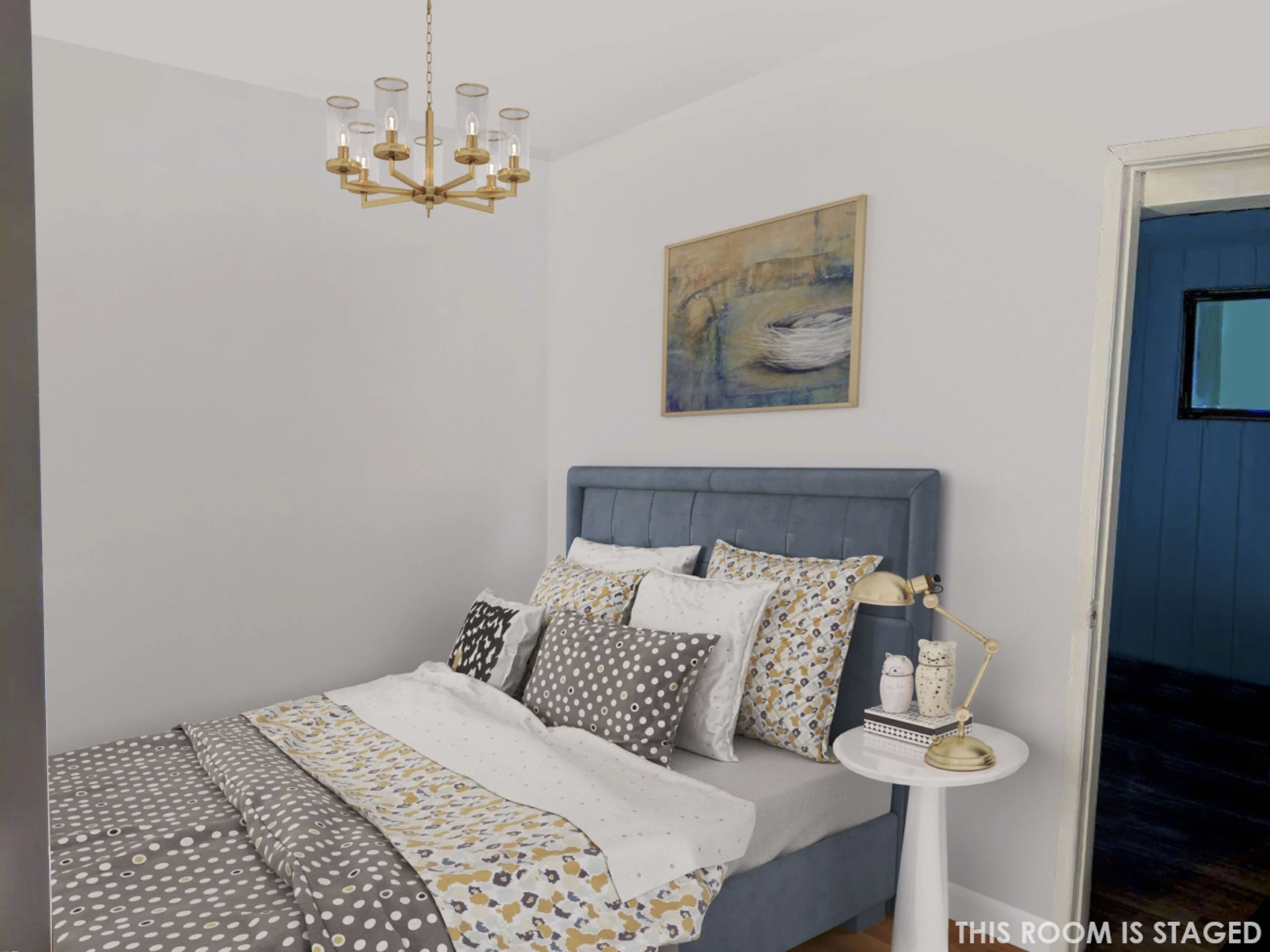A bedroom with a blue upholstered headboard, white walls, and a white side table with a gold desk lamp and decorative jars. A painting hangs above the bed, and a gold chandelier is on the ceiling.
